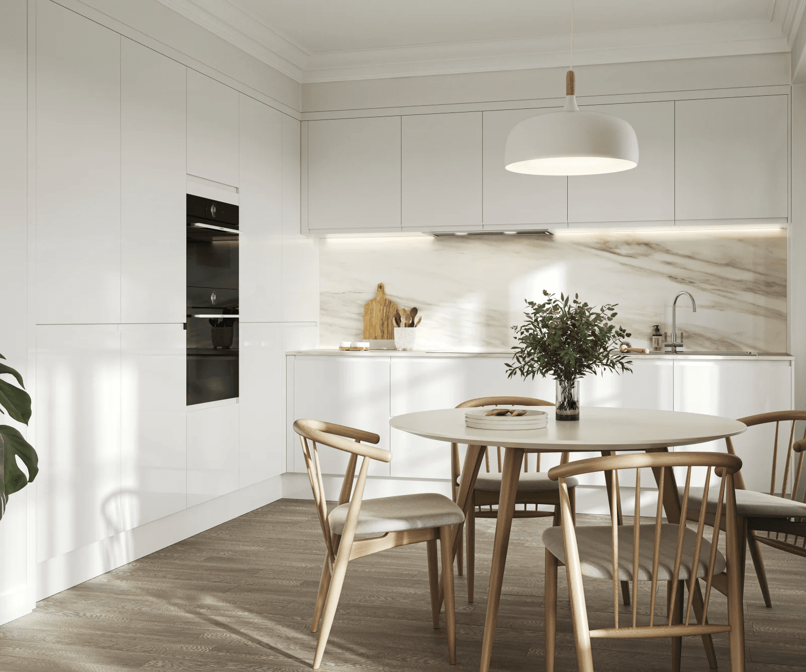The image shows a modern kitchen and dining area with a clean, minimalist design. There are no people visible. The space features white cabinetry with a high gloss finish, a marble-look backsplash, and integrated appliances, including a double oven. A round white dining table with wooden legs is surrounded by four wooden chairs with cushioned seats. On the table, there's a vase with green leafy branches and a small round tray with wooden utensils. A large white pendant lamp hangs above the table, and natural light casts shadows on the hardwood floor.