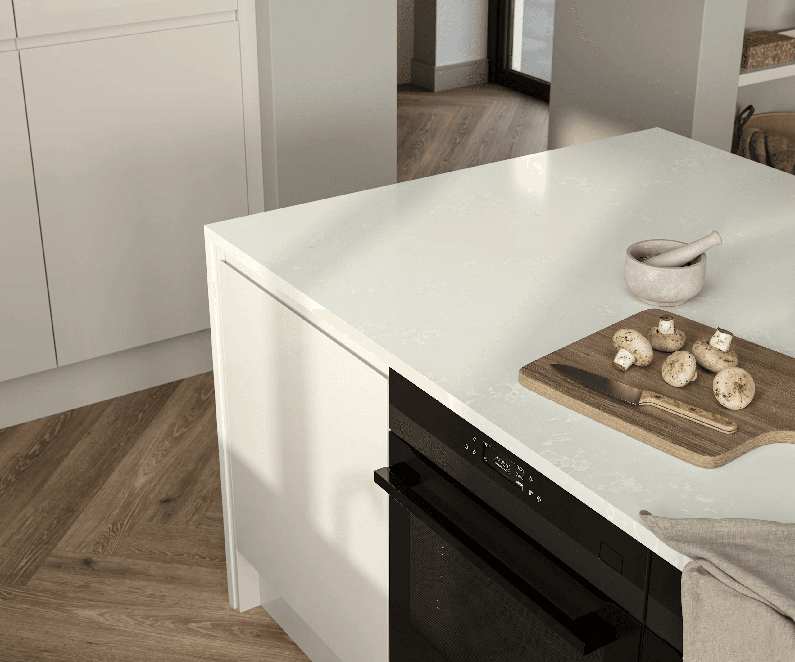 The image shows a close-up view of a modern kitchen island with a white countertop. On the countertop, there is a small wooden cutting board holding five whole mushrooms and a knife with a wooden handle. Next to the cutting board, there is a stone mortar and pestle. The kitchen island features a built-in black oven with a digital display showing a temperature of 225°C. The floor is made of light brown wood arranged in a herringbone pattern. There are white cabinets surrounding the island and part of a glass door is visible in the background.