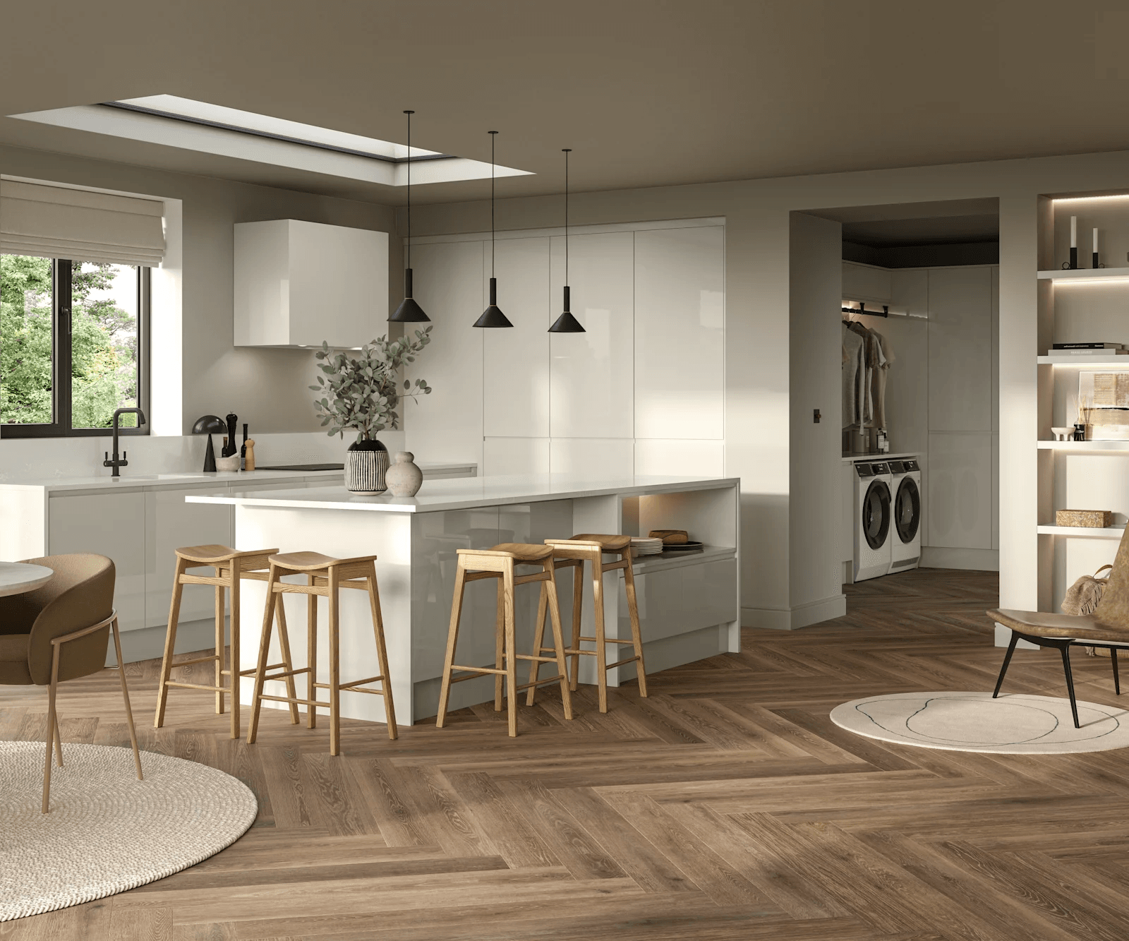 The image depicts a modern, open-plan kitchen and living space with natural light entering through a window and a skylight. The kitchen features glossy white cabinets and a large island with four wooden bar stools. There are three black pendant lights hanging over the island. A vase with branches and two decorative items sit on the island. To the right of the kitchen, there is a laundry area with a washer and dryer and a small hanging clothes rack. The flooring is a herringbone-style wood pattern, extending through the visible space. Seating areas include a brown chair near a round rug and a dark chair near a smaller circular rug. Built-in shelves with decorative objects and books are visible on the right side.