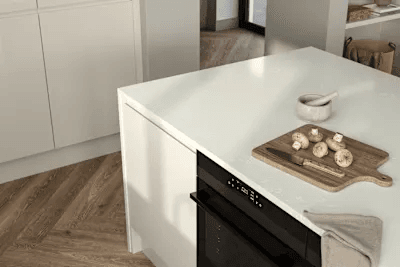The image shows a modern kitchen setting focused on a white kitchen island with a black built-in oven on one side. On top of the island, there is a wooden cutting board with whole mushrooms and a white mortar and pestle. There is also a folded beige kitchen towel hanging on the island's edge. The floor features a light wood herringbone pattern, and white cabinetry is visible in the background.