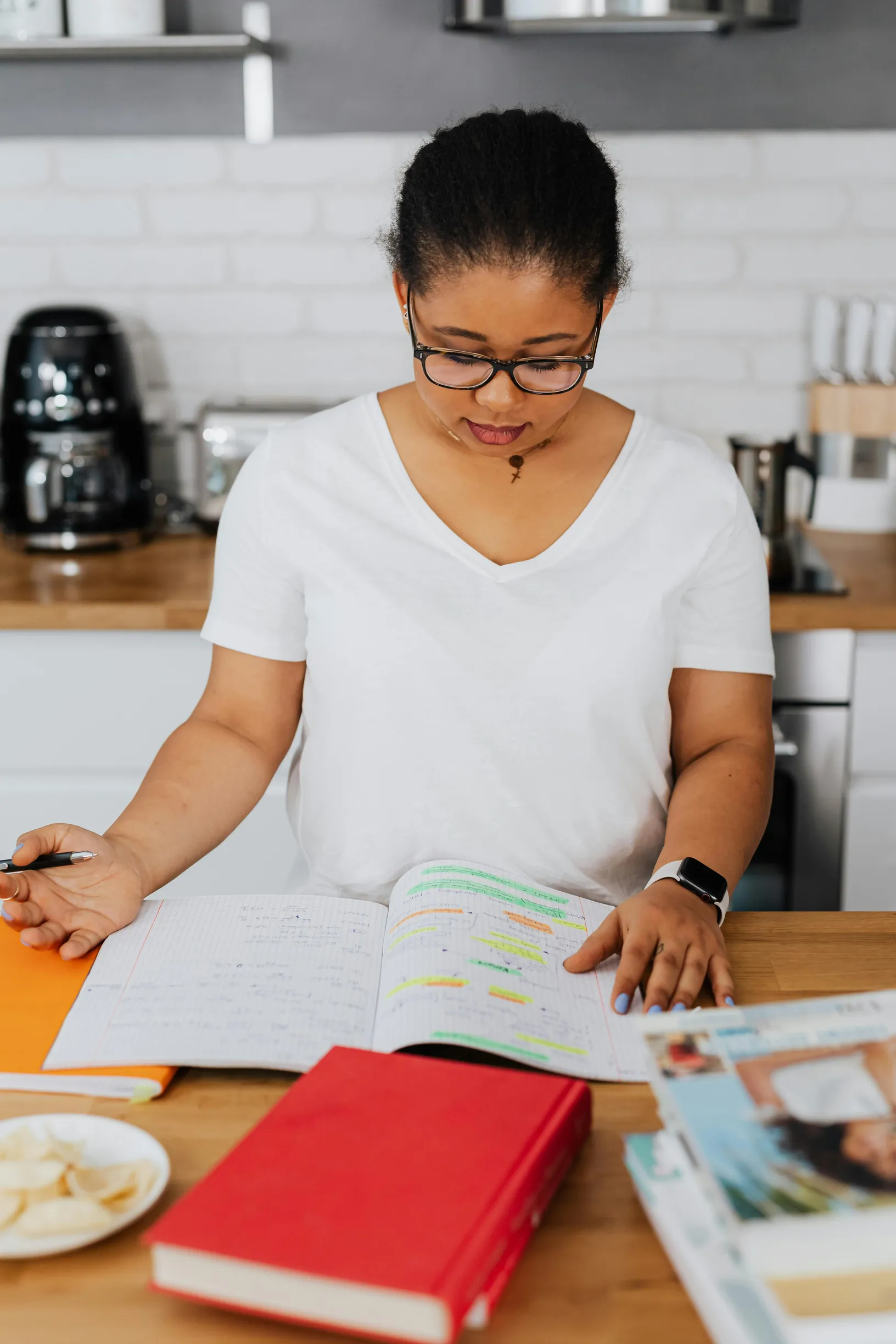 A woman with glasses is seated at a kitchen counter or table, reviewing an open book with highlighted text. She is wearing a white V-neck shirt and has a smartwatch on her left wrist. The setting appears to be a home kitchen with a coffee maker, white tiled backsplash, and wooden countertops visible in the background. The camera angle is eye-level, showing the woman from mid-torso up with a focus on her and the open book. Several other books and papers are on the table in front of her, including a red book 