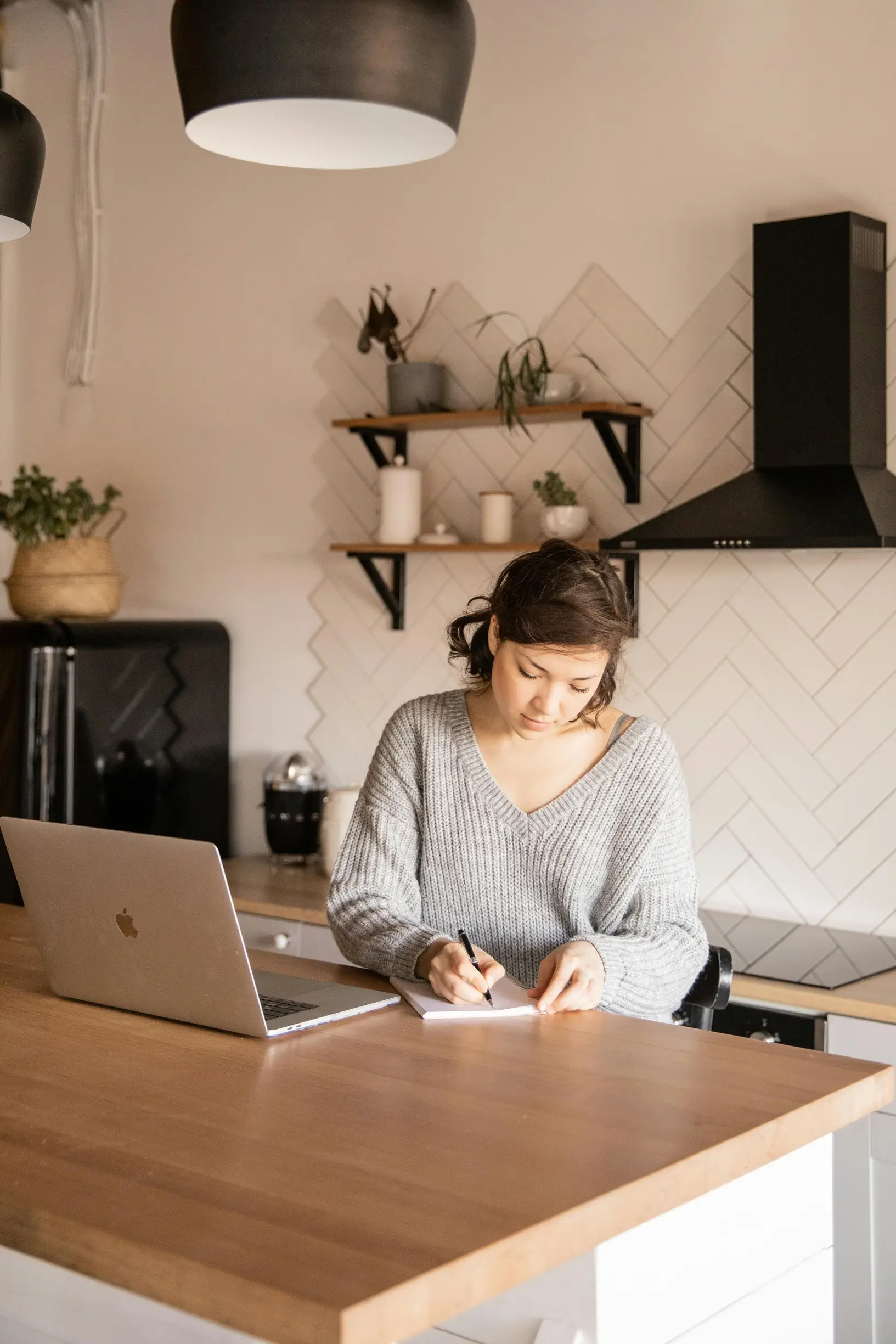 A young woman with medium-length dark hair tied back is seated at a kitchen island counter inside a modern kitchen. She is wearing a light gray knitted sweater and writing in a notebook with a pen. A laptop is open on the counter to her left. The kitchen features a black refrigerator, black range hood, white cabinets, and light wood surfaces. There are two black pendant lights hanging above the counter, and wooden shelves mounted on a white tiled wall hold some small plants and kitchen items.