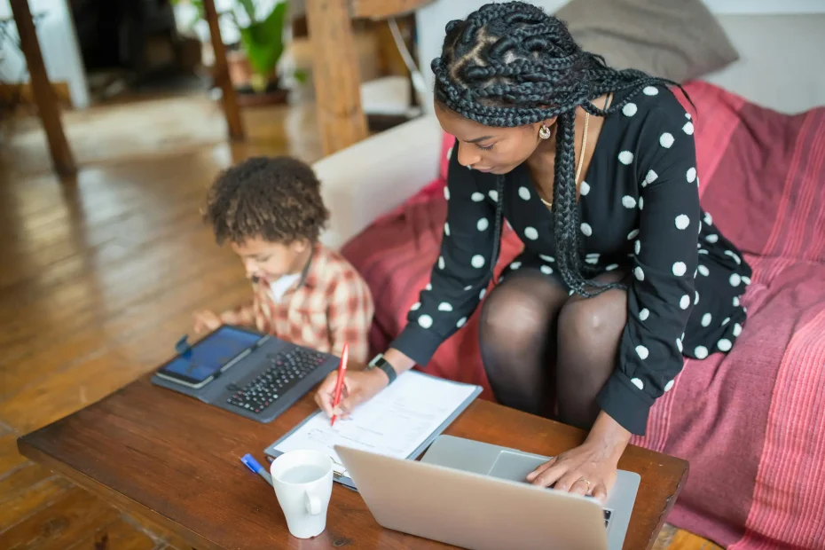 {"description": "A young woman and a child are seated together in a cozy indoor living room setting. The woman, wearing a black dress with white polka dots, is focused on writing in a notebook on a wooden coffee table, while a silver laptop is open in front of her. The child, dressed in a red and white plaid shirt, is sitting next to her, engaged with a tablet device and a keyboard. A white ceramic mug is visible on the table. The room features wooden flooring and furniture, with soft natural light illumina