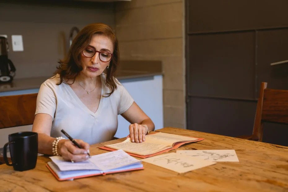 {"description": "An adult woman with light skin and shoulder-length brown hair, wearing glasses and a white or cream short-sleeve V-neck shirt, is seated at a wooden table in an indoor setting. She is writing in a notebook with a pen, with an open book and loose sheets of paper, including sketches, spread out on the table. The background includes a tiled wall and some cabinets or a counter. There is a black mug on the table to her left. The setting appears to be a casual, dimly lit indoor room, possibly a k