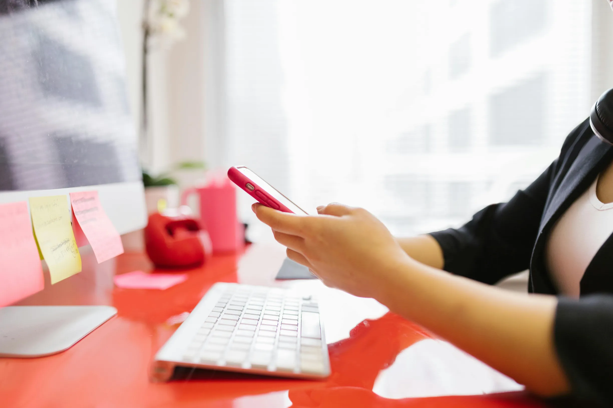 {"description": "The image shows a person seated at a bright red desk using a smartphone with both hands. Only the person's arms and partial torso are visible, wearing a black long-sleeve top over a white undershirt. The setting appears to be a modern indoor office or workspace, characterized by a large window with abundant natural light in the background. On the desk, there is a white keyboard positioned centrally, and a computer monitor edge is partially visible on the left side with several colorful stic