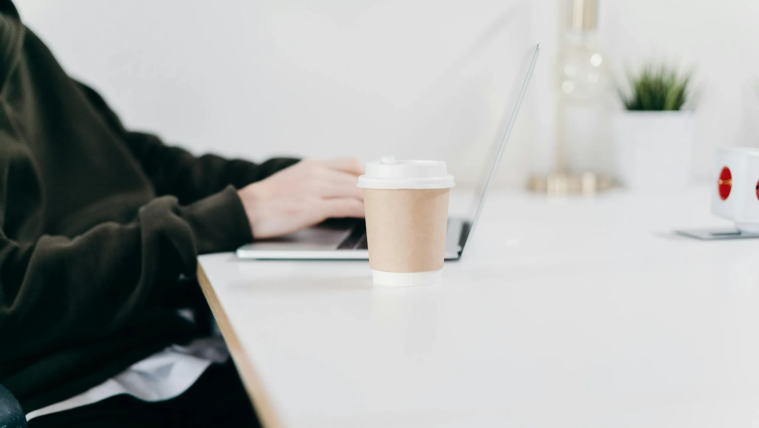 {"description": "The image shows a person seated at a white desk working on a laptop. The person is wearing a black long-sleeve top and a white undershirt, with only their torso and hands visible. On the desk in front of them is a disposable, brown paper coffee cup with a white plastic lid. In the background to the right, a white mug with a red design and a small green plant in a pot are visible, along with a transparent container with a tapered shape. The setting appears to be indoor with soft, diffused li