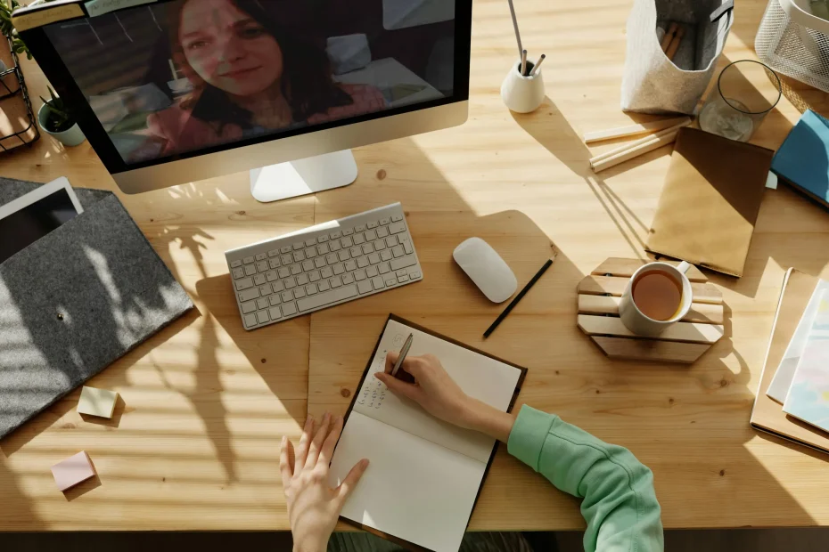 {"description": "The image shows a top-down view of a wooden desk surface in a well-lit indoor setting. A computer monitor displays the face of a woman engaged in a video call. On the desk are a white keyboard and mouse, an open notebook in which a person wearing a green long-sleeve shirt is writing, a pencil, a cup of coffee on a coaster, and various other items including a gray fabric-covered notebook, sticky notes, and some office supplies organized in containers. Sunlight filters onto the desk creating