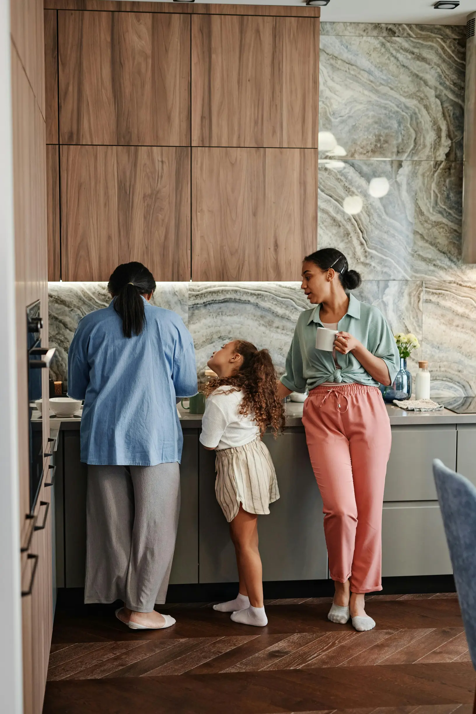 {"description": "The image shows three people in a modern kitchen with a marble backsplash and wooden cabinetry. On the left, an adult facing away from the camera wears a blue top and gray pants, interacting with the countertop, possibly preparing food. In the middle, a young girl with curly hair, dressed in a white sweater and beige skirt with white socks and shoes, looks up towards the adult on the right. On the right, an adult woman wearing a light green top, pink pants, and white socks stands with headp