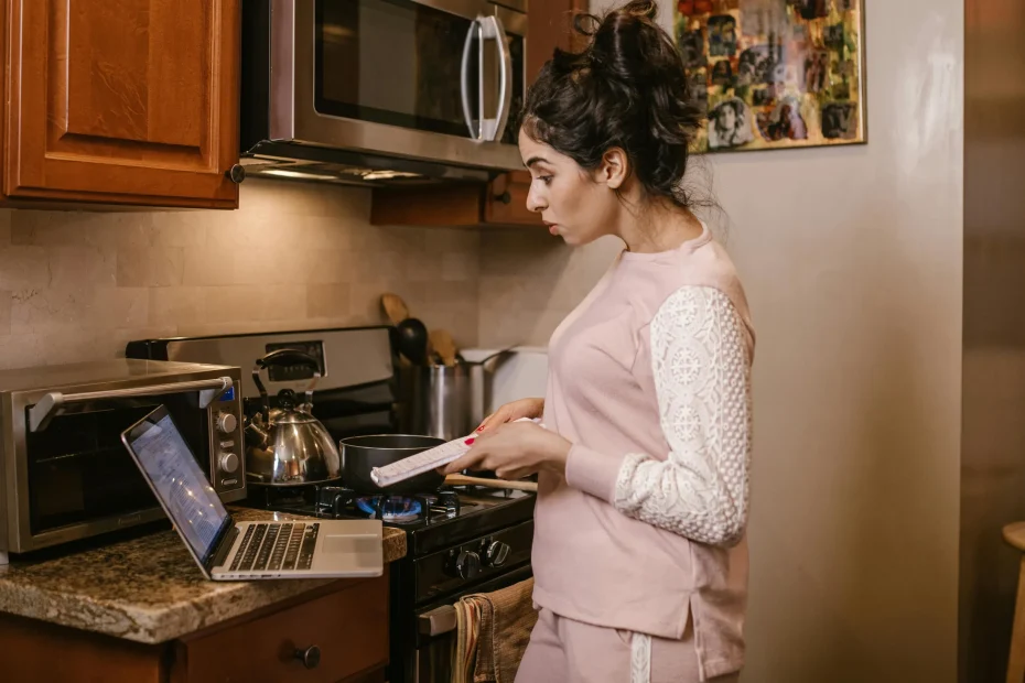 Female multitasking in the kitchen, cooking while using a laptop for online learning.
