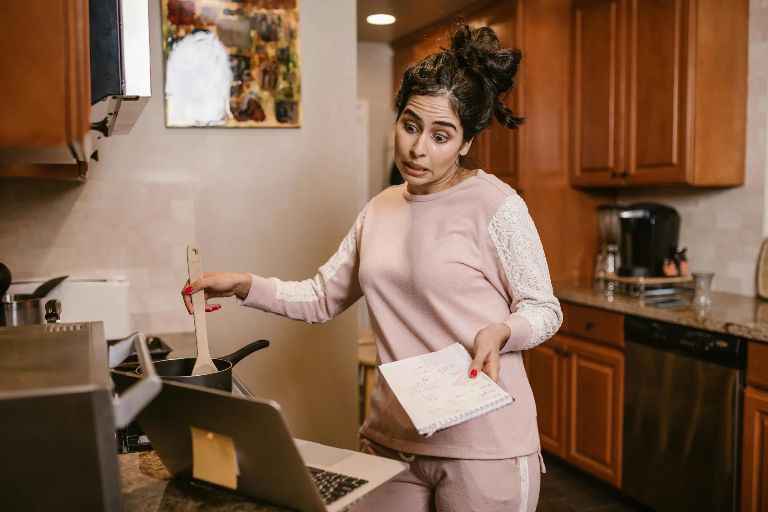 {"image_loaded": true, "load_issue": null, "description": "A woman stands in a domestic kitchen, stirring a pot on the stove with a wooden spoon while looking at a laptop on the counter. She holds a notebook or recipe book in her other hand. The kitchen features brown wooden cabinets, a light-colored backsplash, and granite countertops. There is a coffee maker and other kitchen utensils visible on the counter. The lighting is warm and even, suggesting artificial indoor lights.", "people": {"count": 1, "role