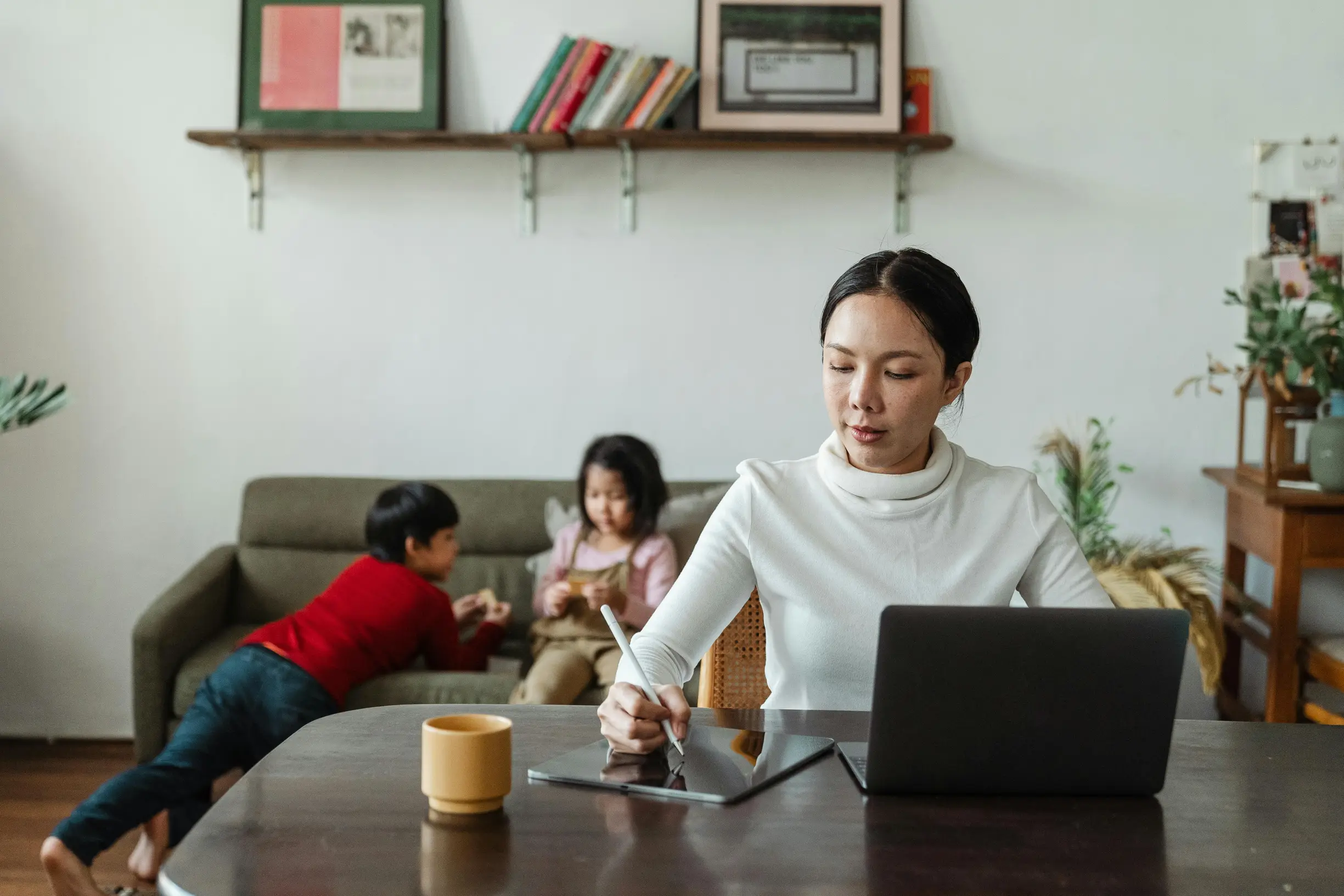 {"image_loaded": true, "load_issue": null, "description": "An adult woman is seated at a wooden table in a home environment, working on a digital tablet with a laptop open in front of her. Behind her on a couch, two young children are engaged with a toy or object. The room features a simple, light-colored wall with a wooden shelf holding books and framed pictures. There is also a houseplant on the right, and a yellow mug on the table near the woman.", "people": {"count": 3, "roles": ["adult working", "child