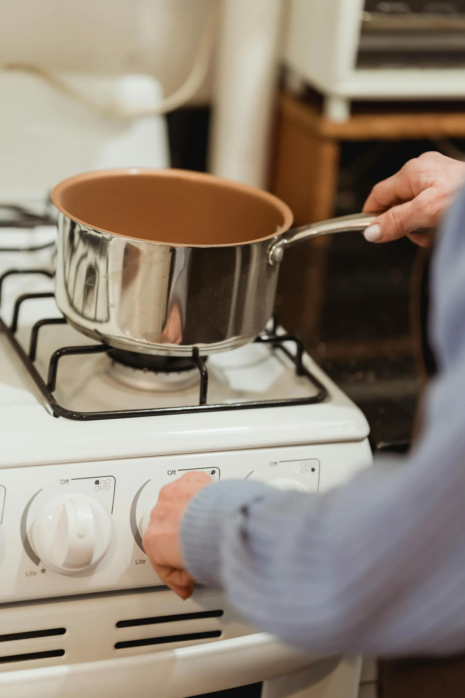 {"image_loaded": true, "load_issue": null, "description": "The image shows a close-up view of a person cooking indoors on a gas stove. The person is adjusting a control knob on the stove with one hand, while holding a stainless steel saucepan with a brown interior with the other hand positioned above the burner. The individual's arm and part of the torso are visible, wearing a long-sleeved, light blue textured garment. The stove is white with control dials on the front panel. In the background, kitchen furn