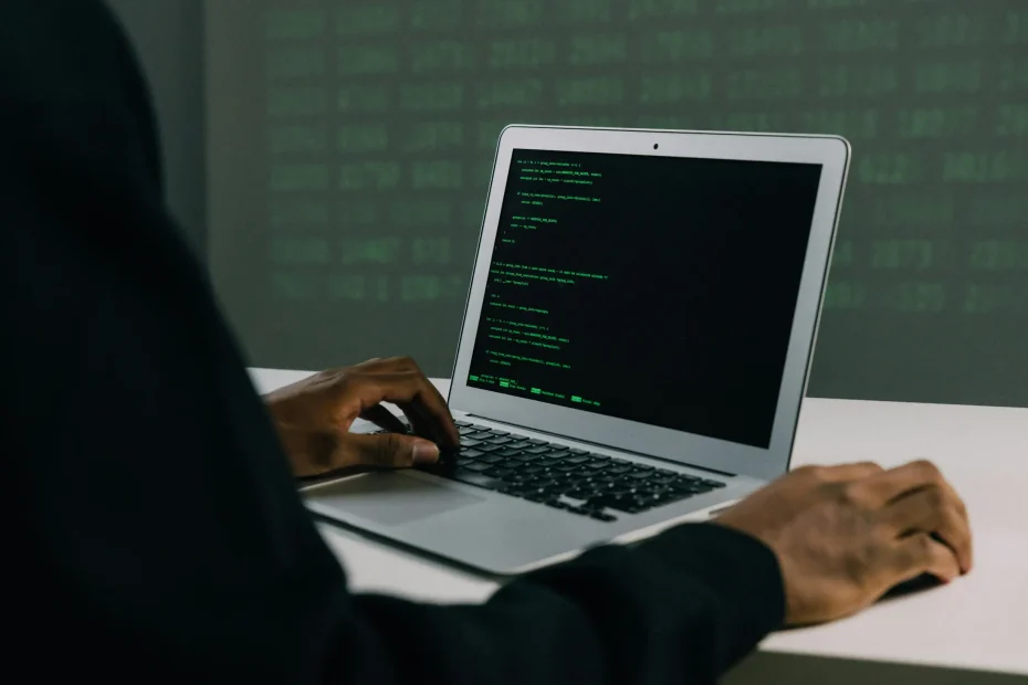 {"image_loaded": true, "load_issue": null, "description": "The image shows a laptop on a white table with code displayed on its screen in green text against a black background. A person with dark skin is seated at the table, their hands positioned near the laptop keyboard and trackpad. The person is dressed in a dark long-sleeve garment, and only their torso and arms are visible from behind and the side. The background is out of focus but shows a wall with faint green text or code. The camera angle is eye-l