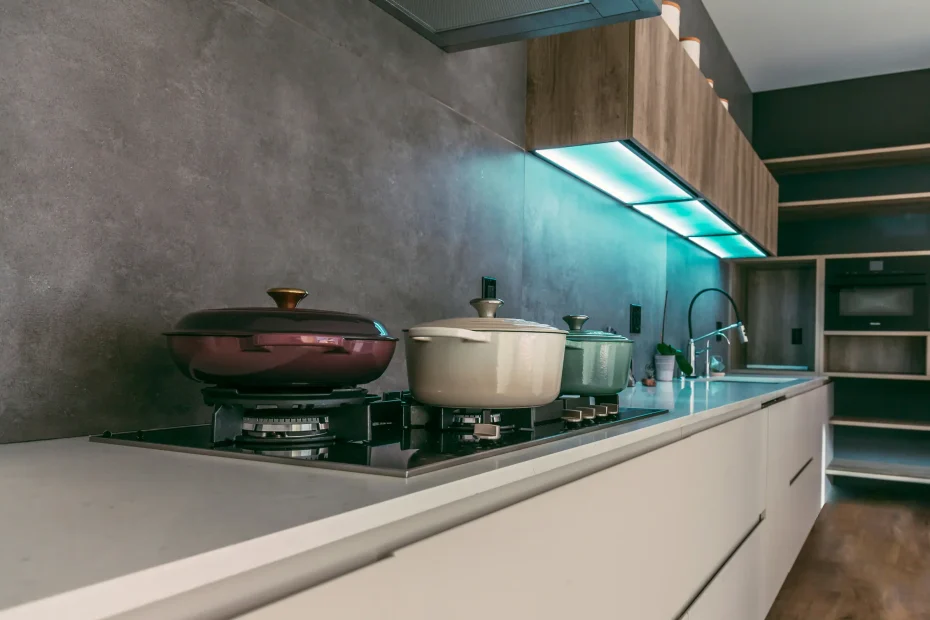 {"image_loaded": true, "load_issue": null, "description": "The image shows a modern kitchen countertop with a built-in gas stove. There are three pots with lids on the stove: one maroon, one cream-colored, and one light green. Above the stove, there is a set of wooden cabinets with under-cabinet lighting emitting a blueish glow. The countertop is a smooth light gray or white surface, and the backsplash is a matte, textured dark gray material. To the right, there is a sink with a modern faucet and some small