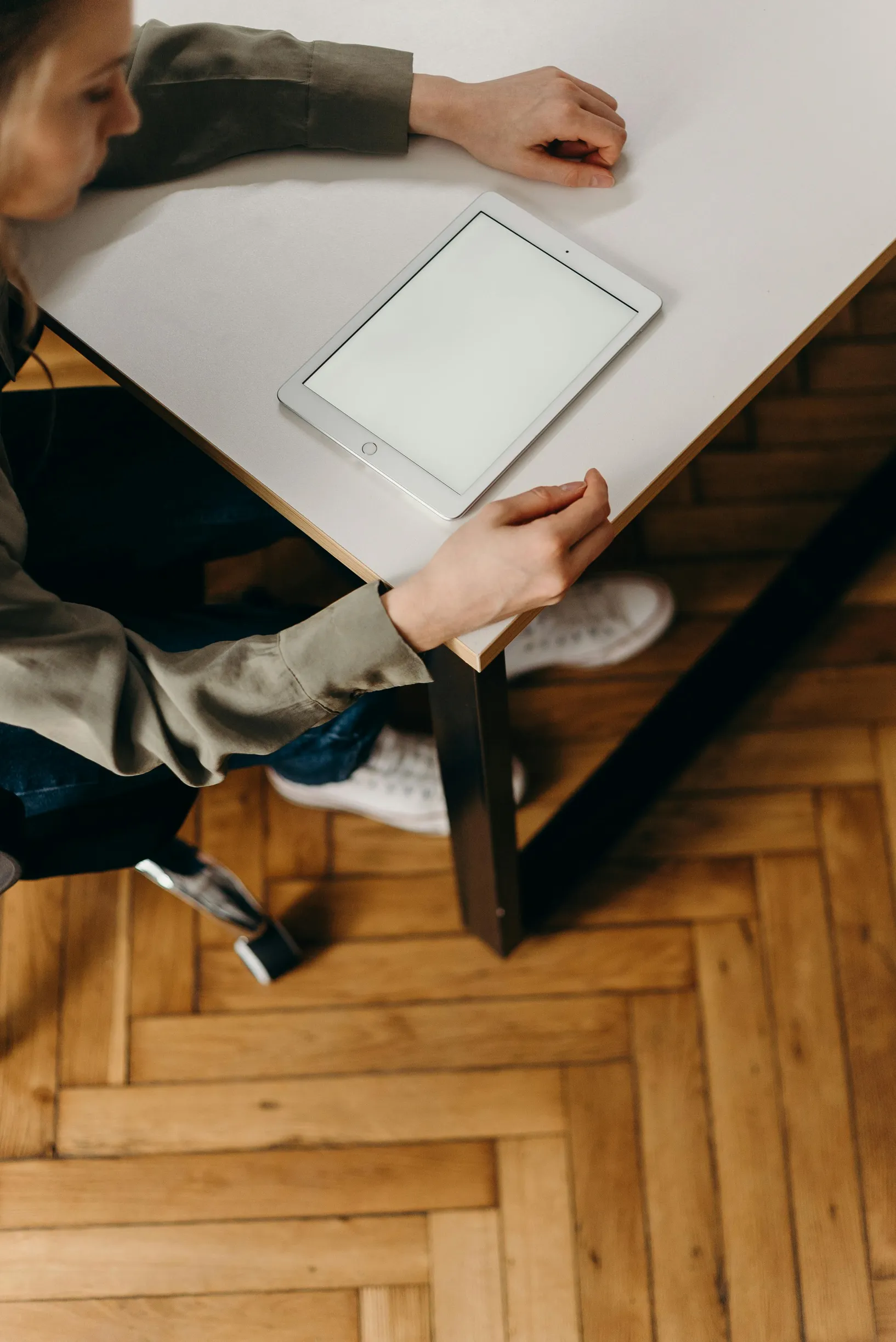 {"image_loaded": true, "load_issue": null, "description": "The image shows a person seated at a wooden parquet floor in front of a white table. The person is viewed from above and slightly to the side, with their hands resting on the table near a white tablet device placed on the surface. The person's clothing includes a greenish long-sleeve shirt and dark pants. One white sneaker is visible on the floor. The setting appears to be indoors with warm lighting. The photo is taken from an elevated angle, captur