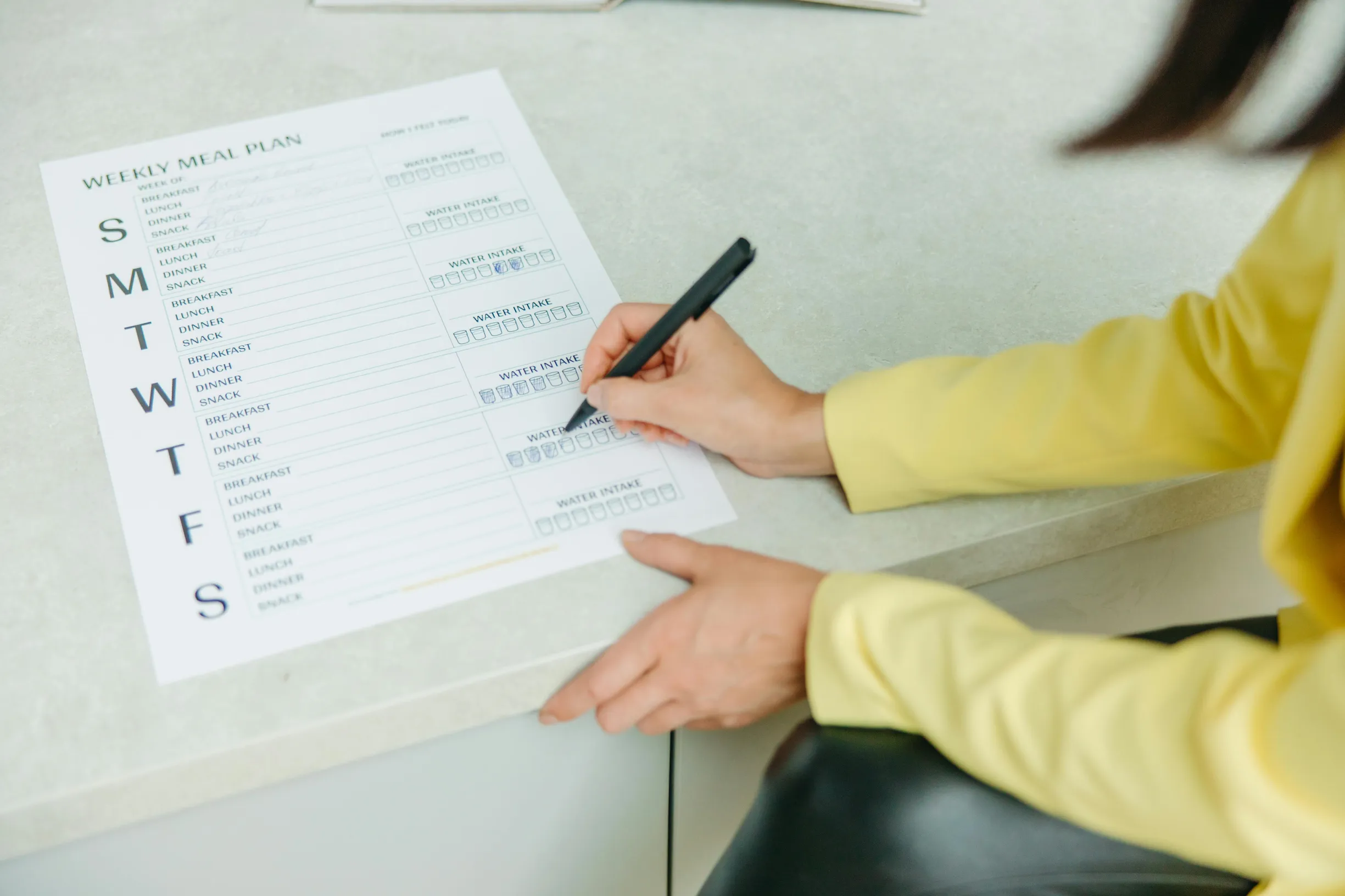 {"image_loaded": true, "load_issue": null, "description": "The image shows a person sitting at a light-colored countertop, filling out a printed weekly meal plan sheet with a black pen. The person is wearing a yellow long-sleeve top and black pants, and only their torso and hands are visible. The meal plan sheet is organized by days of the week with columns for each day, featuring text and empty boxes for writing. The lighting is bright and natural or artificial. The camera angle is a slight overhead perspe