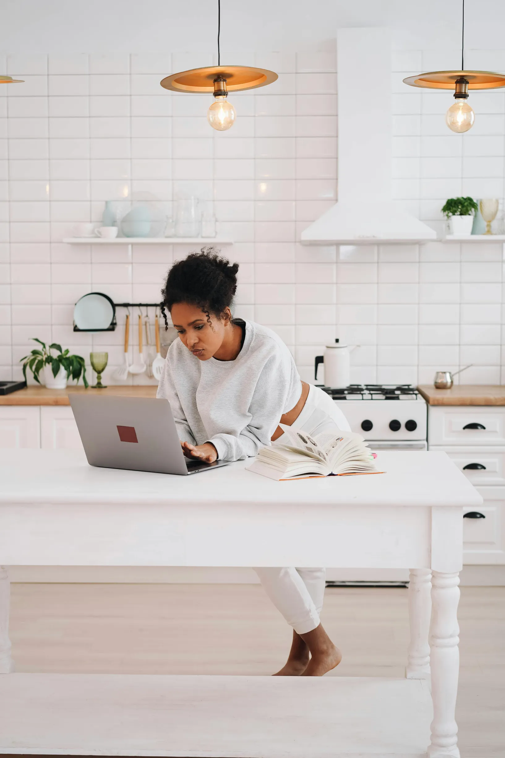 {"image_loaded": true, "load_issue": null, "description": "The image shows a single person, an adult woman with dark curly hair, in a bright, modern kitchen. She leans on a white kitchen island while working on a laptop. The kitchen features white tiled backsplash, white cabinetry, and wooden countertops. Various kitchen items, including plants, cooking utensils, and a kettle, are visible, creating a clean and organized environment. The lighting is bright and natural, enhancing the white and neutral tones t