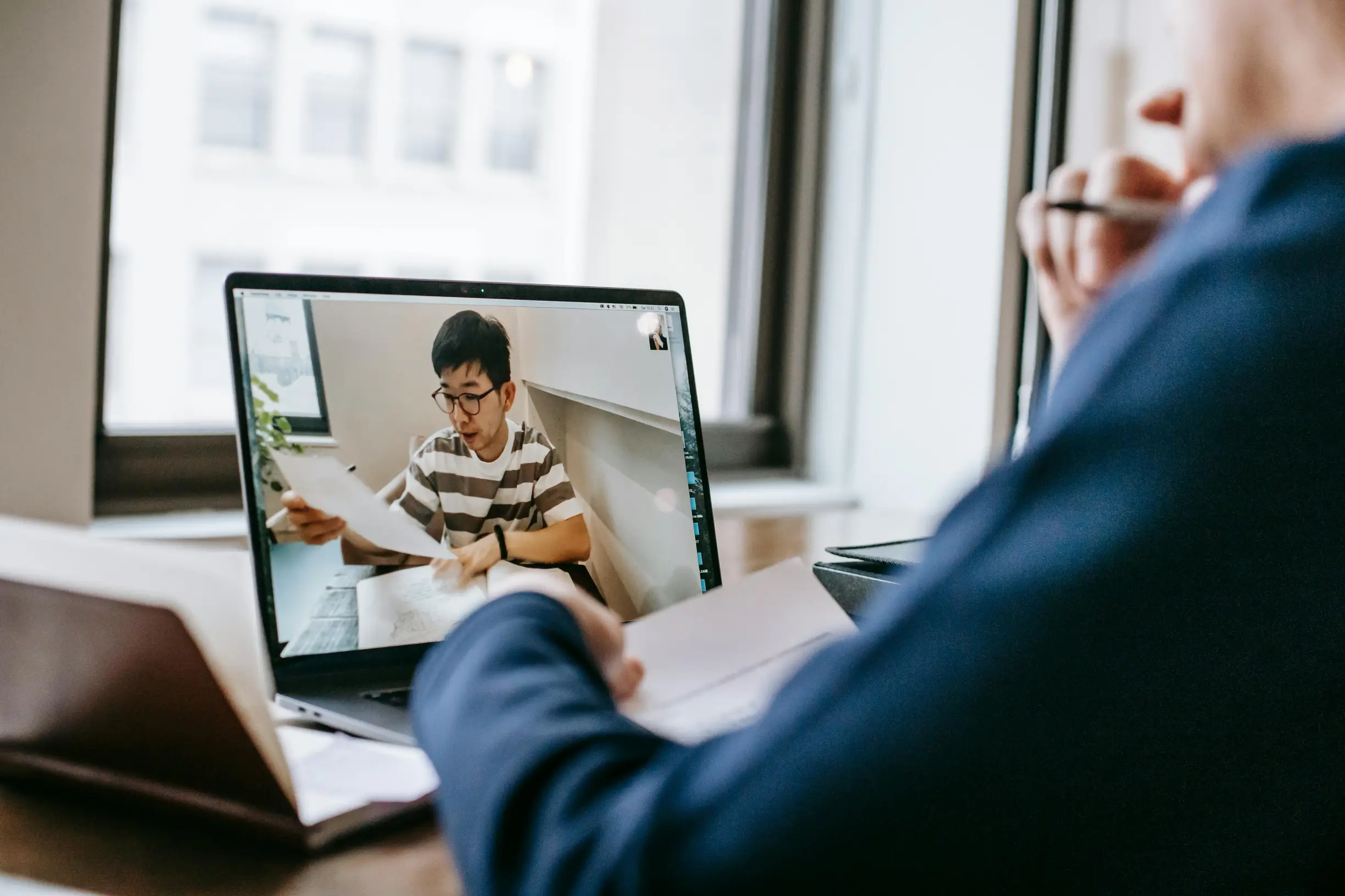 The image shows a person sitting at a desk in front of a laptop engaged in a video call with another person visible on the laptop screen. The person on the screen appears to be a young male with short dark hair and glasses, wearing a striped t-shirt and holding papers. The person at the desk is partially visible from behind, wearing a dark blue long sleeve top and holding a pen and papers. The setting is indoors near large windows with natural light illuminating the scene. The desk has some objects includin