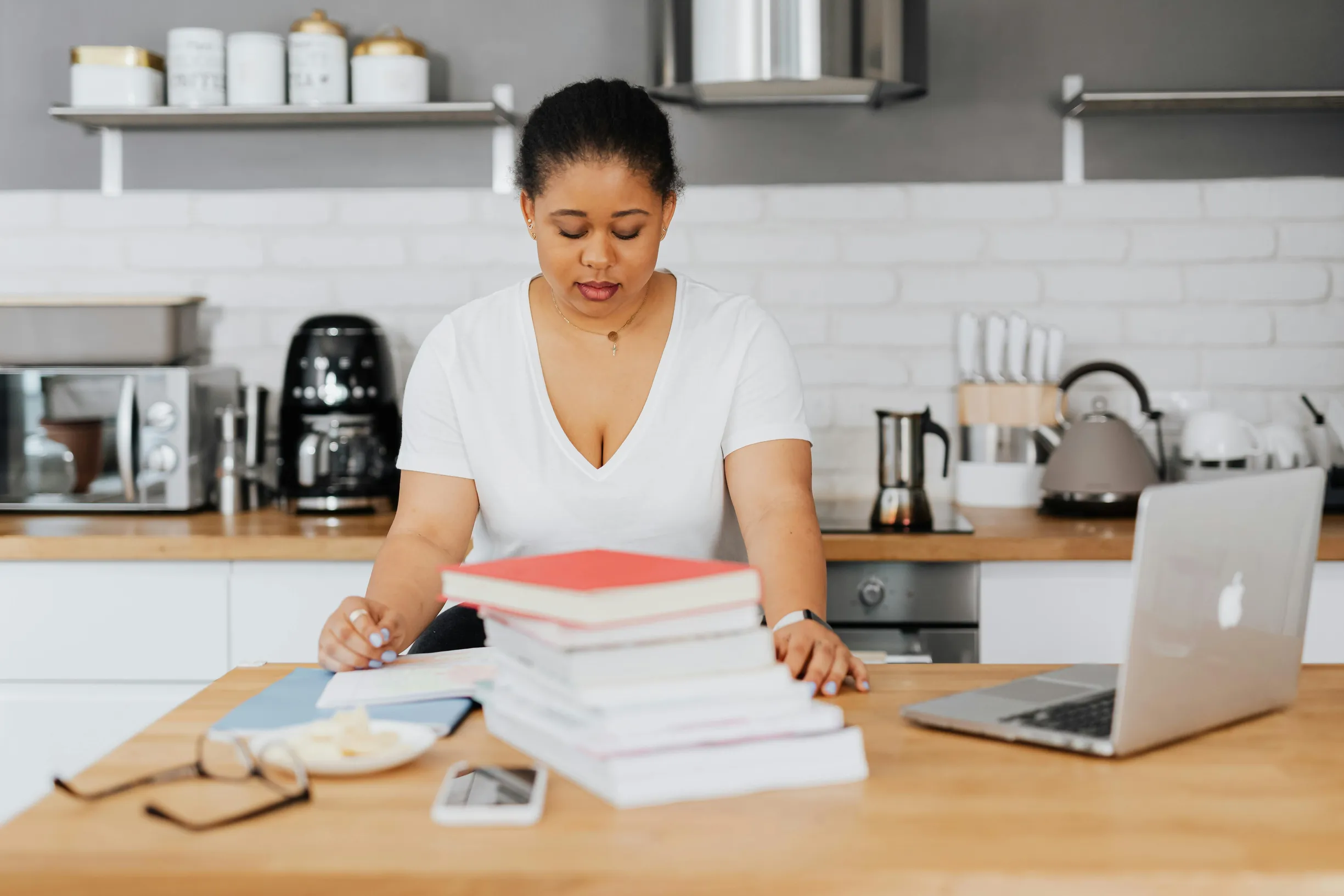 The image shows a woman seated at a kitchen counter or workspace. She is looking down at papers in front of her, with a stack of books or notebooks directly on the table. To her right is an open laptop. The background shows kitchen appliances such as a coffee maker, kettle, toaster, and jars on shelves.