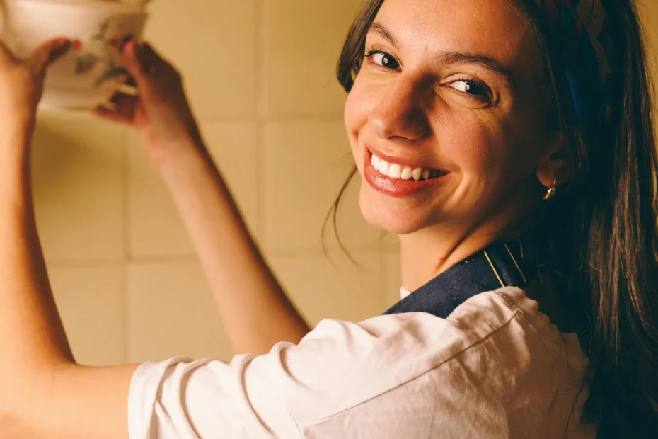 Woman in a cozy kitchen reaching for a ceramic lid, smiling warmly.
