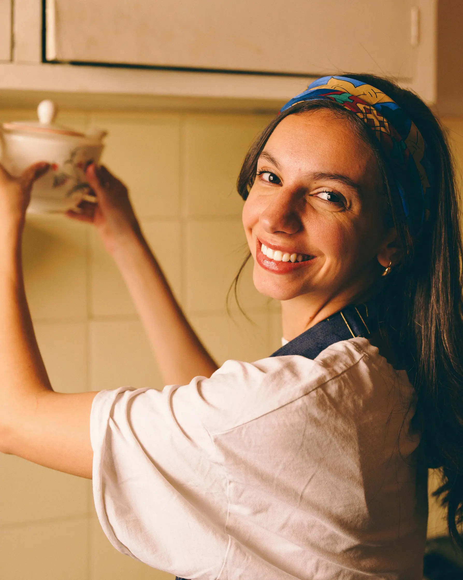 Woman in a cozy kitchen reaching for a ceramic lid, smiling warmly.