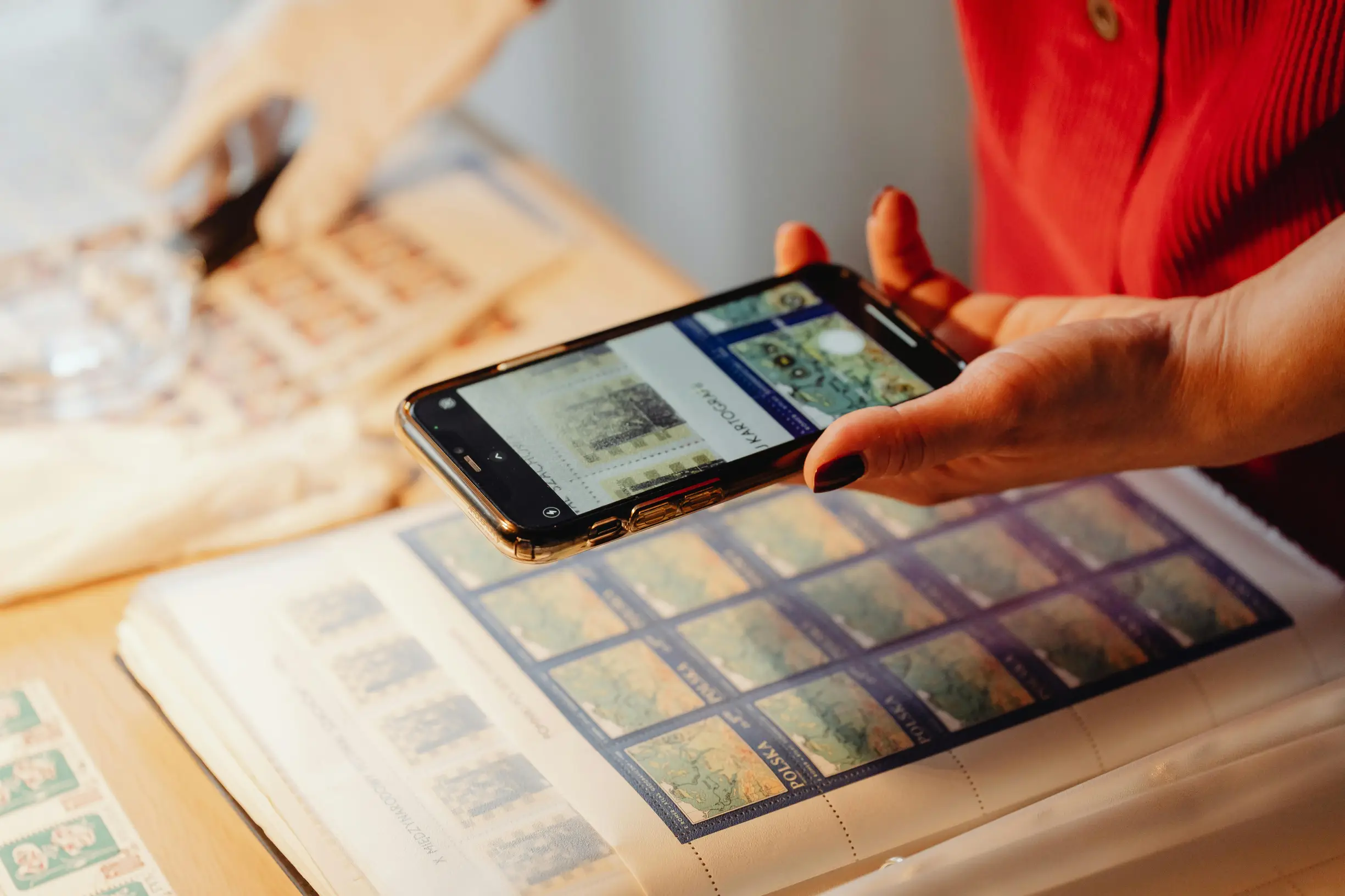A close-up image showing two hands engaged in an activity involving a smartphone and a stamp collection album. One hand holds a smartphone, displaying images of stamps on its screen, while the other hand is slightly blurred in the background, appearing to interact with the stamp album on a wooden table.