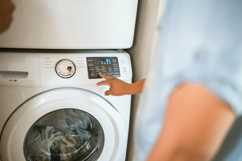 A close-up image shows a front-loading washing machine with clothes visible inside through the circular door window. An adult person's hand is adjusting the control panel on the upper right section of the machine, which displays digital settings and buttons. The person is partially visible from the side, slightly out of focus, wearing a light blue short-sleeve shirt. The background appears to be an indoor laundry room with neutral-colored walls.