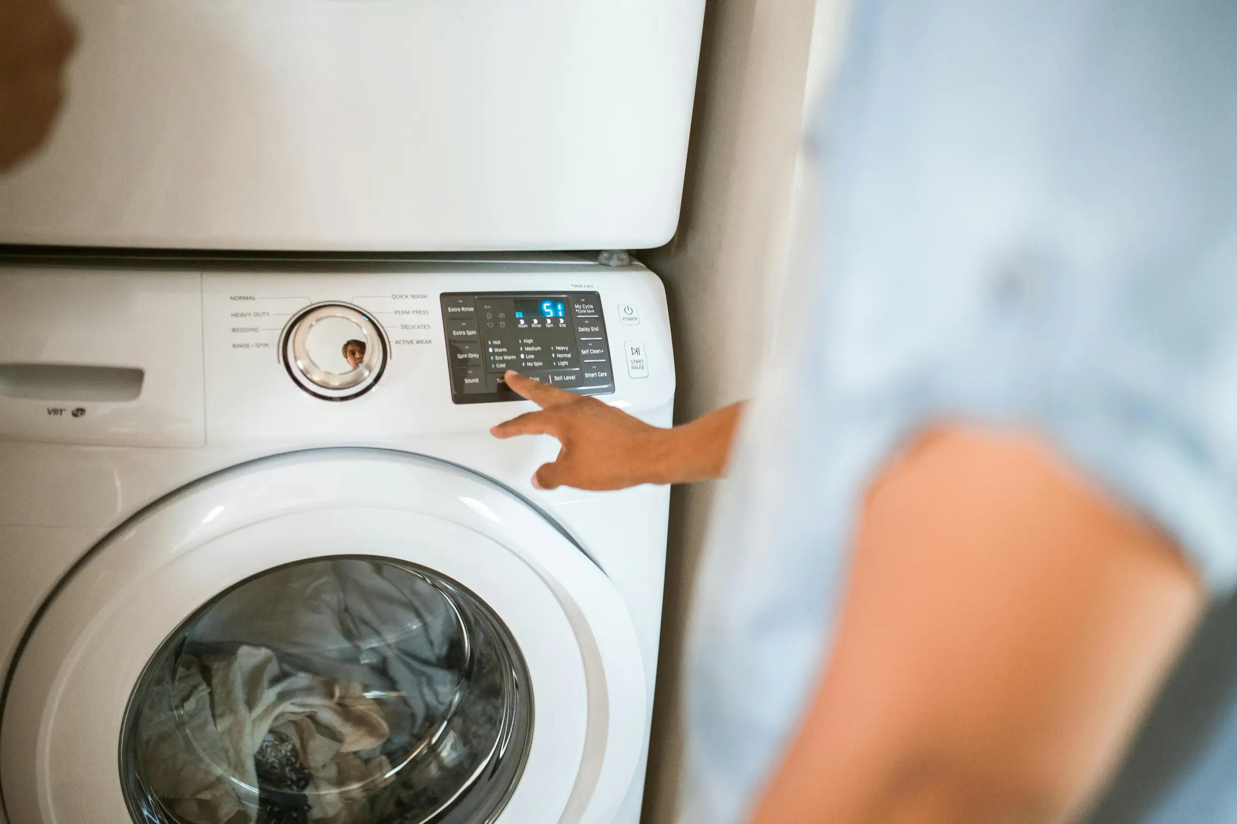 A close-up image shows a front-loading washing machine with clothes visible inside through the circular door window. An adult person's hand is adjusting the control panel on the upper right section of the machine, which displays digital settings and buttons. The person is partially visible from the side, slightly out of focus, wearing a light blue short-sleeve shirt. The background appears to be an indoor laundry room with neutral-colored walls.