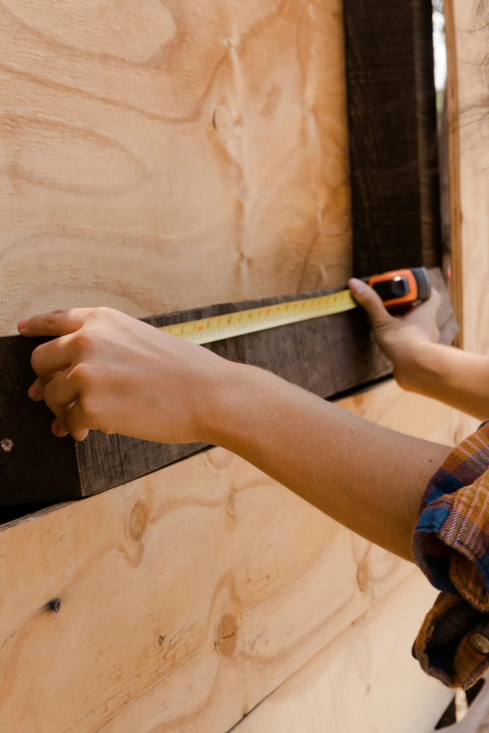 The image shows a close-up of a person's arms and hands measuring a piece of dark wood against a light-colored plywood surface using a tape measure.