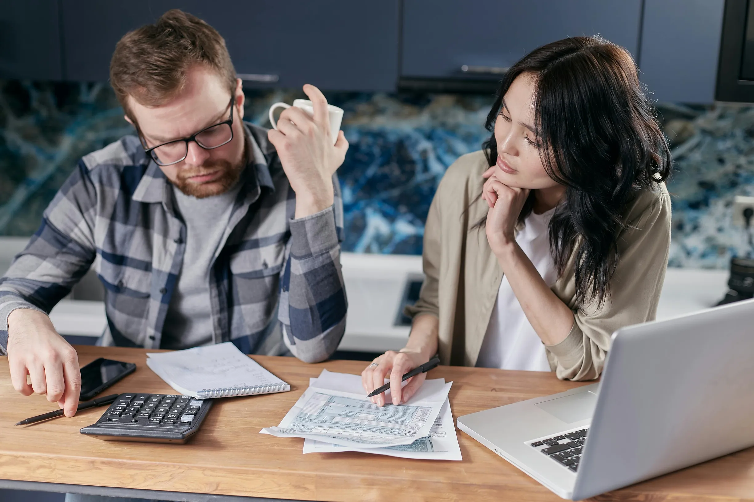 A man and a woman are seated at a wooden table indoors, working together on paperwork. The man, wearing glasses and a plaid shirt over a gray T-shirt, holds a white mug in his right hand while using a calculator with his left. The woman, dressed in a beige jacket over a white shirt, looks attentively at the documents in front of them, resting her chin on her left hand and holding a pen with her right hand. On the table are scattered papers, a notebook, a calculator, a pen, and a silver laptop, suggesting a 
