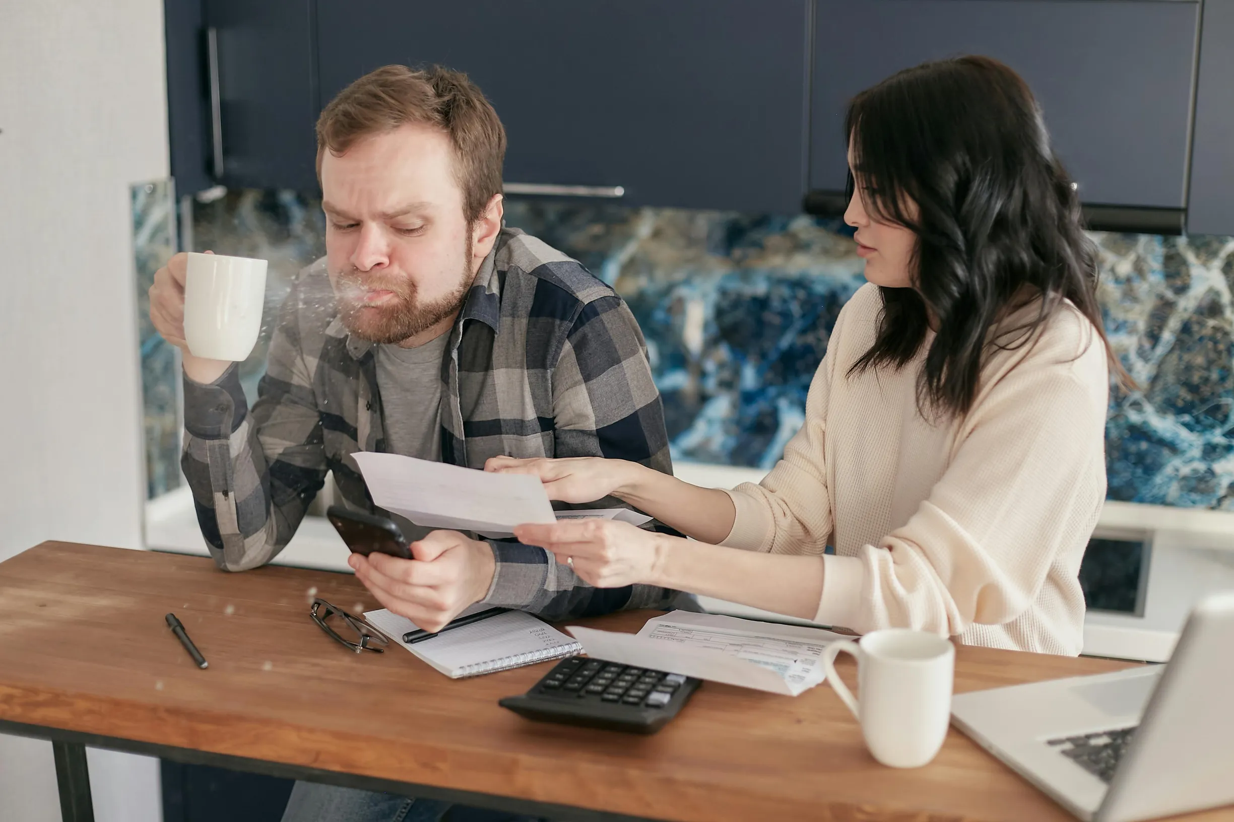 The image shows a man and a woman sitting at a wooden table in a modern kitchen setting. The man is on the left, wearing a black and gray plaid shirt over a gray T-shirt, holding a white mug in one hand and a smartphone in the other while blowing out hot steam or liquid from the mug. The woman on the right is wearing a cream-colored sweater and is holding a piece of paper, appearing to be engaged in discussing or showing the document to the man. On the table are additional papers, a black calculator, a small notebook, a pen, a pair of eyeglasses, a white mug, and a laptop that is partially in frame. The background consists of dark blue cabinetry and a blue and white marbled backsplash. The scene is lit with natural or soft artificial lighting, with a medium framing that captures the upper half of the individuals and most of the table.
