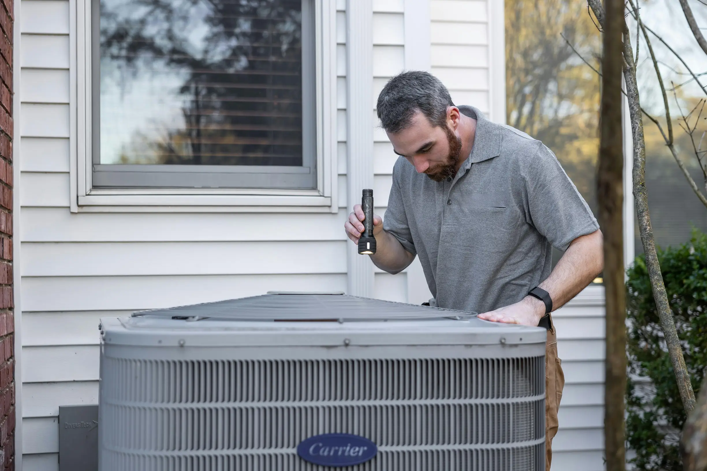 A man is inspecting or repairing an outdoor air conditioning unit labeled 'Carrier' against the exterior of a house with white siding. He is holding a flashlight in his right hand, shining light on the top of the unit, and resting his left hand on it. The setting is outdoors during daytime or early evening. The man is casually dressed in a short-sleeved gray polo shirt and brown pants, with a watch on his left wrist. The background includes a window with a reflection of trees and part of a tree trunk on the right side of the image. The camera angle is eye-level and medium distance, capturing the man from waist up and the AC unit in full.