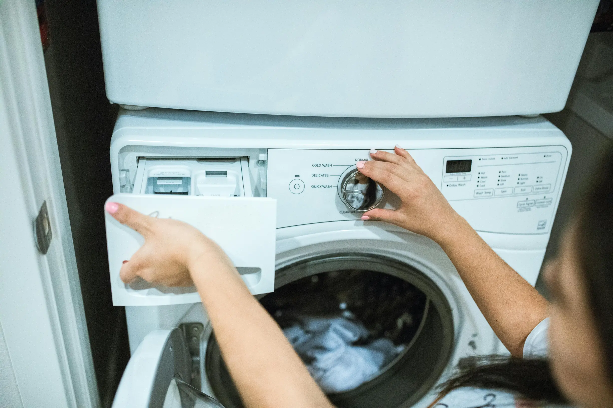 A person is operating a front-loading washing machine indoors. The person's hand is turning the control knob on the washing machine, while the other hand is opening the detergent drawer. The washing machine door is open, revealing some clothes inside. The setting appears to be a laundry area or room with the washer placed under a white appliance possibly a dryer.