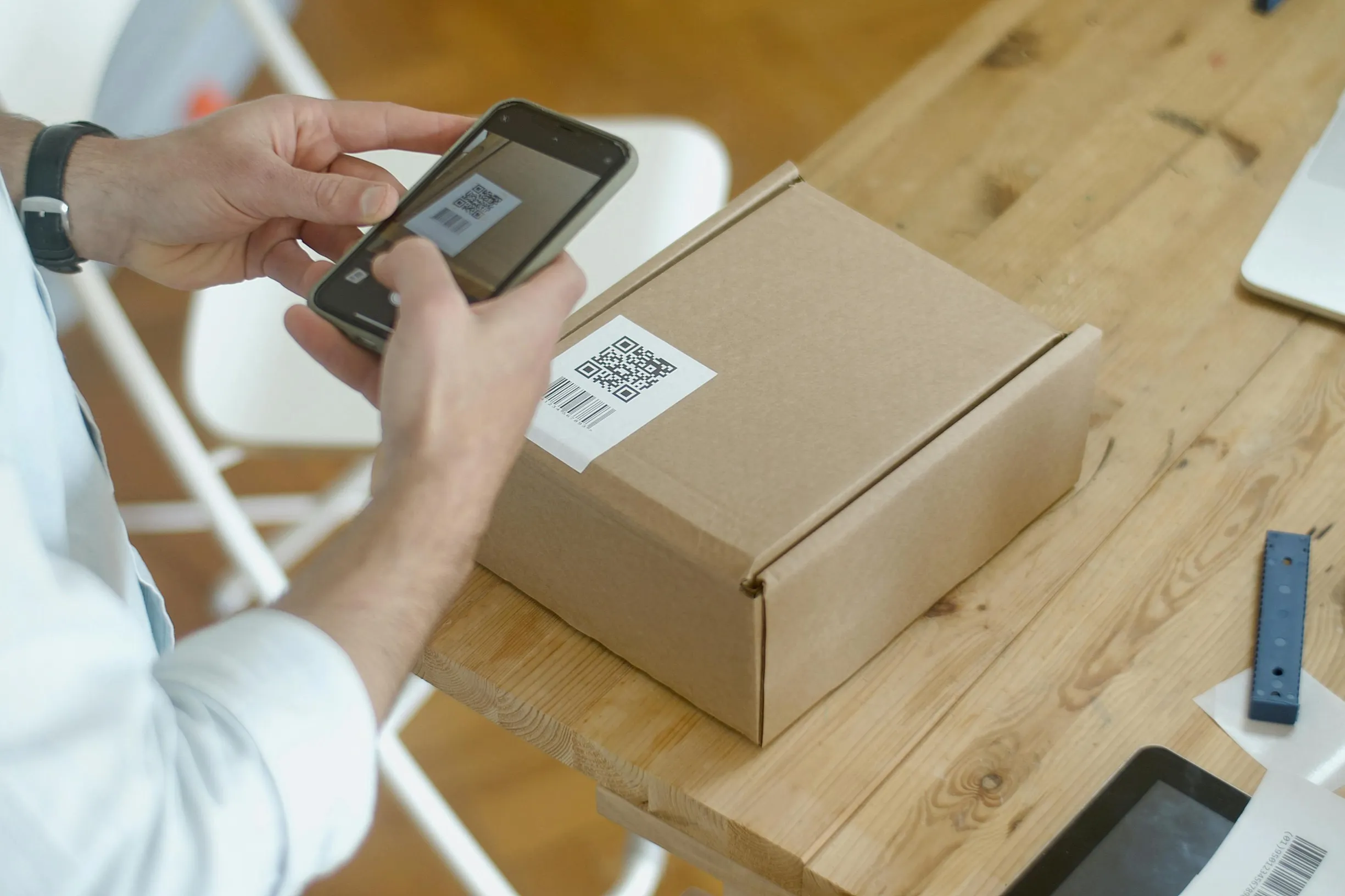 A person is taking a photo of a cardboard box on a wooden table using a smartphone. The box has a white label with a QR code on it. The person's hands are visible, wearing a white long-sleeve shirt and a black watch. On the wooden table, there are a few other objects including a tablet and a small black device. The setting appears to be indoors with a warm, natural lighting and a close-up camera angle focusing on the hands, phone, and box.