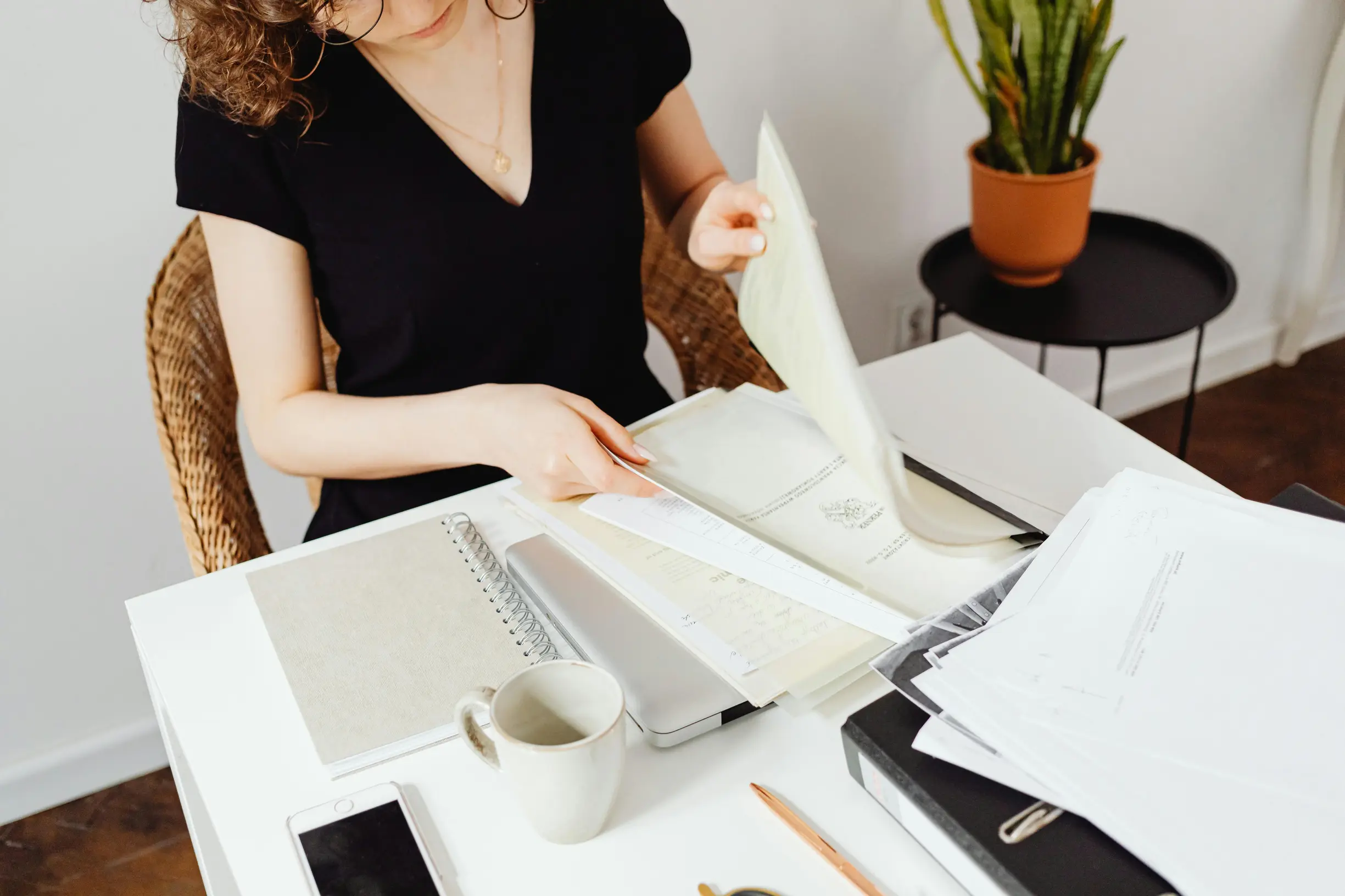 A person with light skin and curly brown hair, wearing a black short-sleeve shirt, is seated at a white desk in a room. They are flipping through several sheets of paper held in their hands. The desk contains various objects including a closed laptop, a gray notebook, a ceramic mug, a smartphone, a wooden pencil, a pen, and a stack of papers organized in black folders. Behind the desk, there is a small black round table with a potted snake plant on top. The room appears indoors with white walls and a wooden