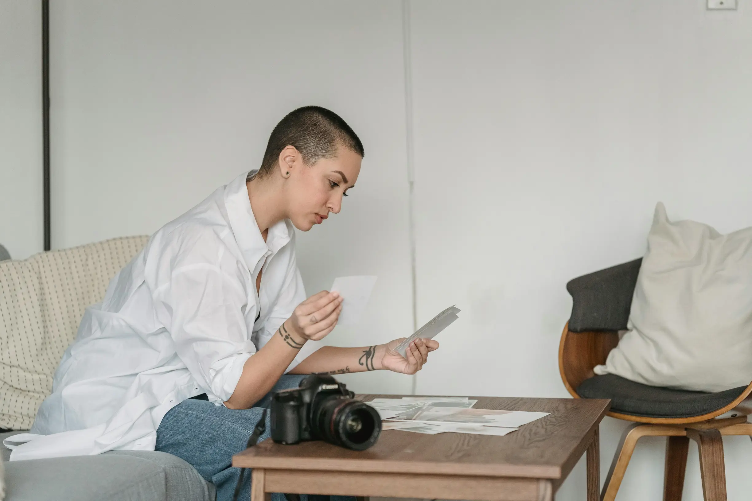 A person with short hair, wearing a loose white shirt and blue jeans, sits on a couch in a minimalist indoor setting. They are examining two photographs or papers in their hands while seated at a wooden table with more papers spread on it and a camera placed on the table. The room has a neutral color palette with white walls, a light gray couch, a wooden chair with cushions in the background, and soft, natural lighting.