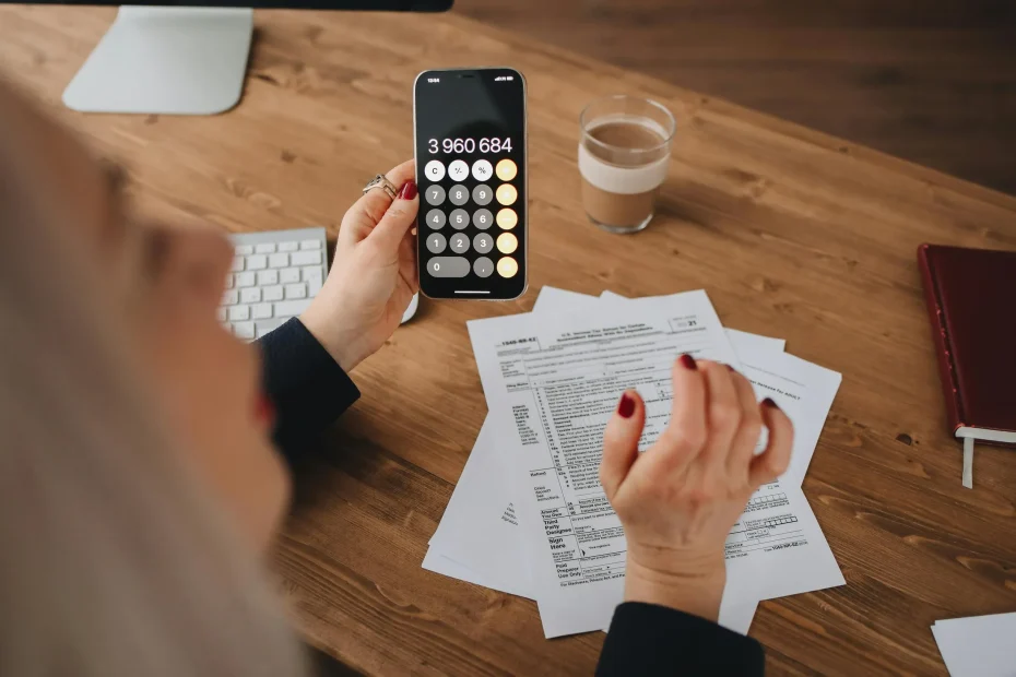 A woman calculates on her smartphone while working with documents at a desk.