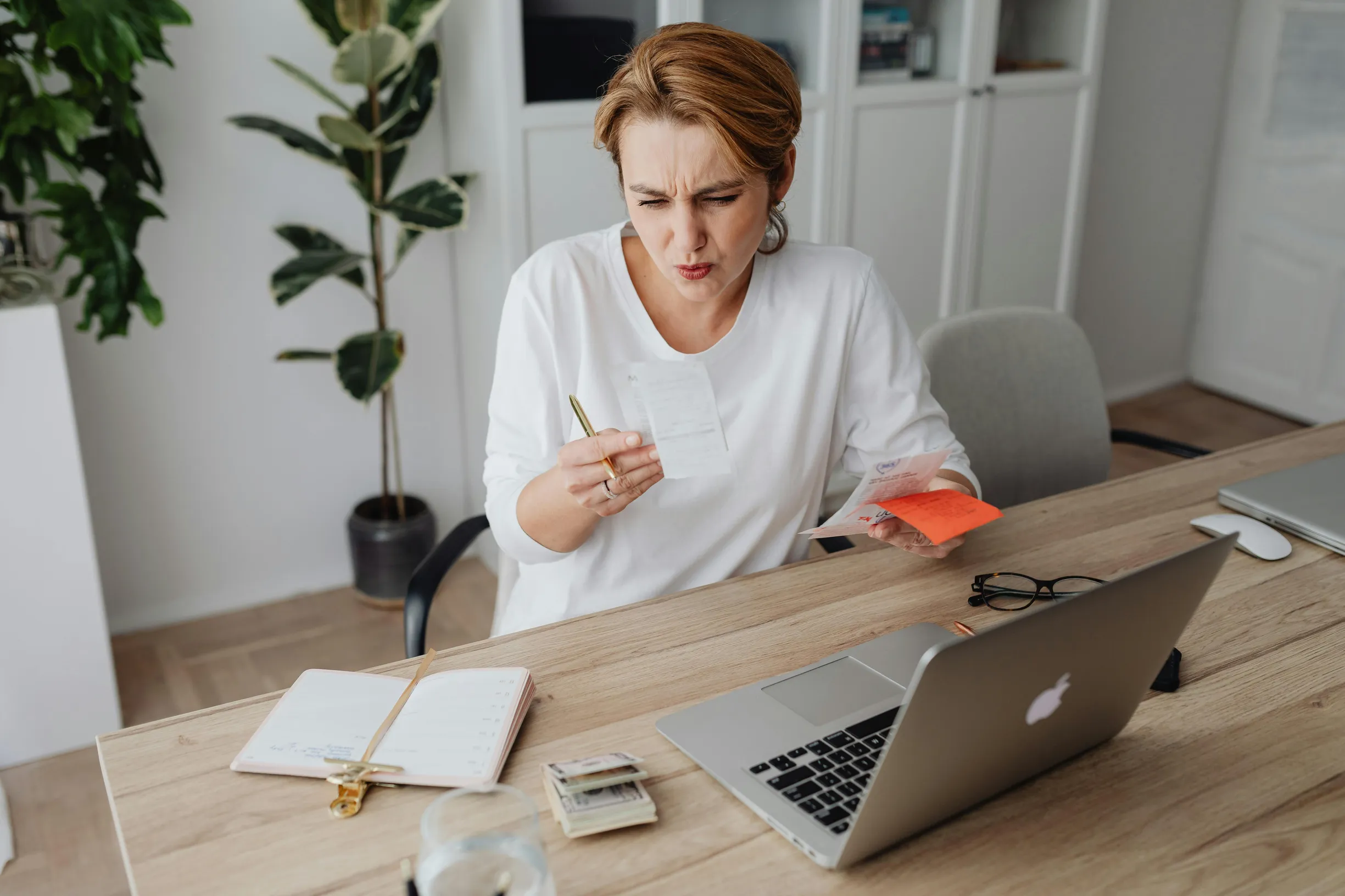 A woman is seated indoors at a wooden desk, looking intently at a receipt in her hand while holding an orange card in the other hand. She appears to be middle-aged with light skin and reddish hair tied back. On the desk are a laptop with an Apple logo, a glass of water, an open notebook with a pen, a pair of eyeglasses, and a few small items. Behind her, there is a white bookshelf and a tall potted plant. The room has light-colored flooring and white walls, illuminated with natural or diffused light. The ca