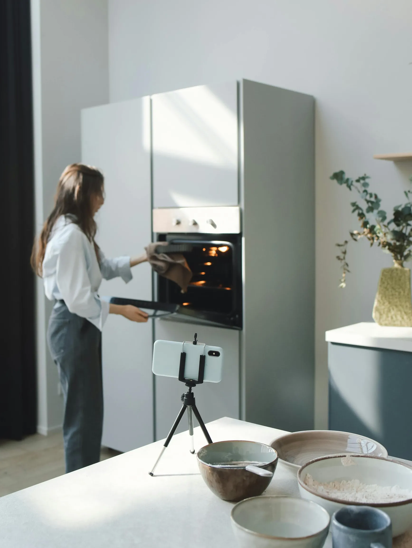A woman is seen from the side placing or removing food from a built-in oven in a modern kitchen. The kitchen has a minimalistic design with light-colored cabinetry and an open layout. A smartphone is mounted on a small tripod on a counter in the foreground, facing the woman. Several ceramic bowls and plates are arranged on the kitchen island or countertop.