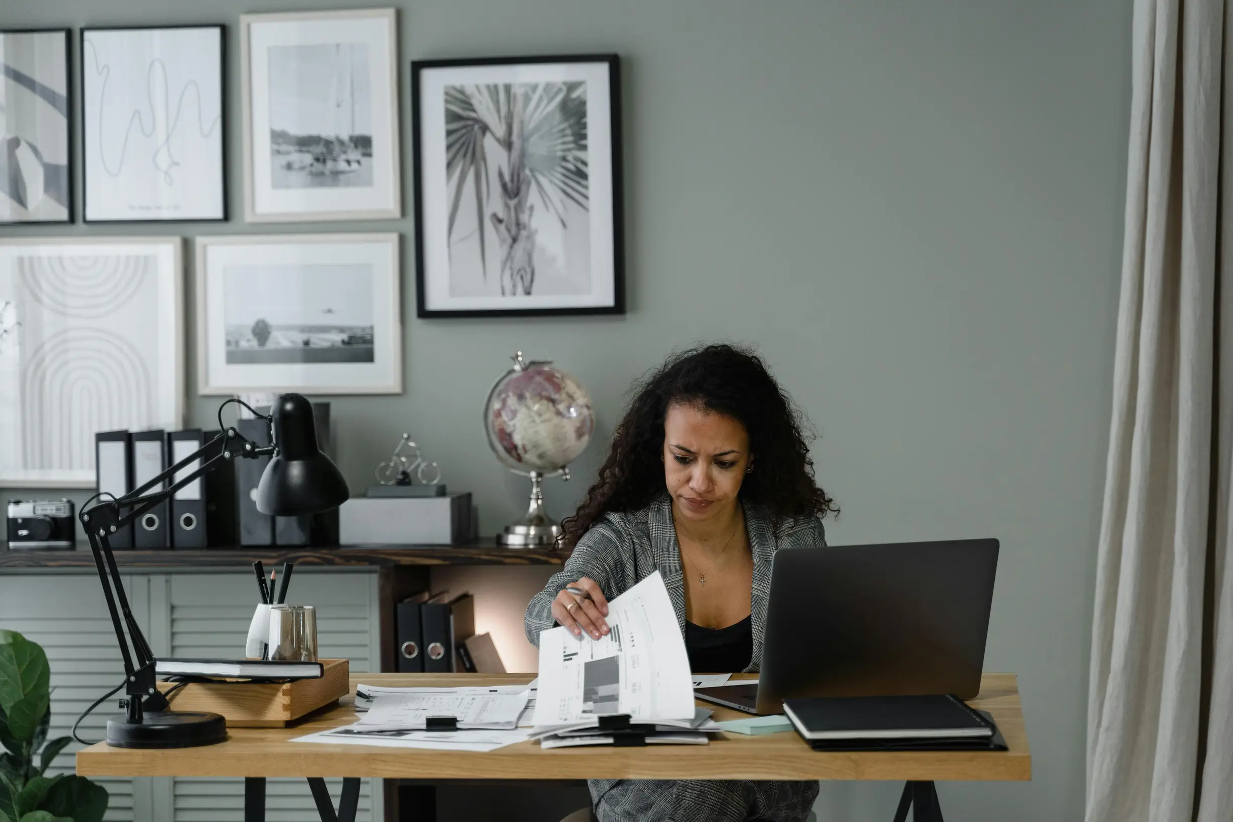 A woman sits at a wooden desk in a modern, minimalistic office space. She is examining papers in front of her, surrounded by office supplies, a laptop, and a globe on a shelf behind her. The wall features several framed black-and-white artworks, and a black desk lamp is positioned on the left side of the desk. The lighting appears natural and evenly distributed, with the photo taken from an eye-level angle showing a medium framing of the scene.