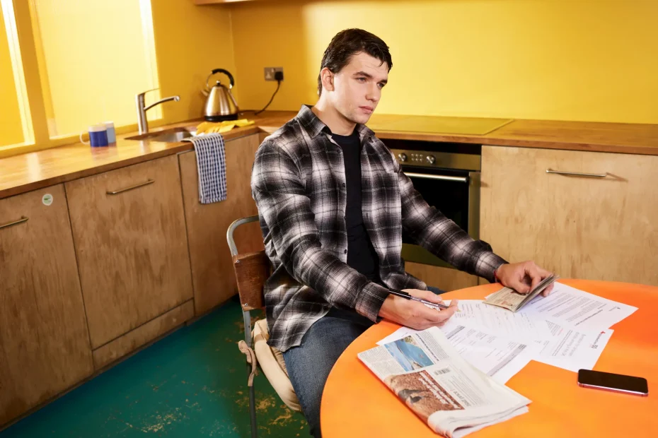 A young adult man with short, dark hair sits at a round orange table in a kitchen setting. He is wearing a black and white plaid shirt over a black t-shirt and appears focused on signing or filling out papers spread out on the table. The kitchen has wooden cabinets with silver handles, a yellow wall, green floor, and a window letting in natural light. Visible objects include a kettle on the countertop, a blue mug near the window, a smartphone on the table, and an assortment of documents and a newspaper.