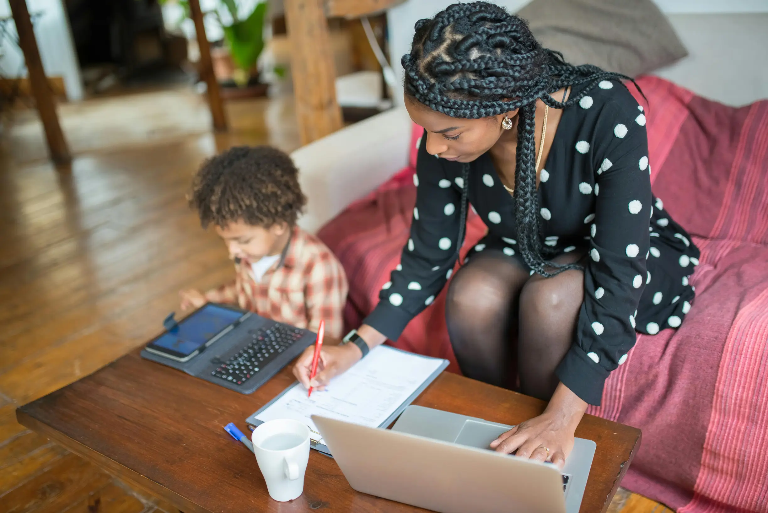 A young woman and a child are seated together in a cozy indoor living room setting. The woman, wearing a black dress with white polka dots, is focused on writing in a notebook on a wooden coffee table, while a silver laptop is open in front of her. The child, dressed in a red and white plaid shirt, is sitting next to her, engaged with a tablet device and a keyboard. A white ceramic mug is visible on the table. The room features wooden flooring and furniture, with soft natural light illuminating the scene, c