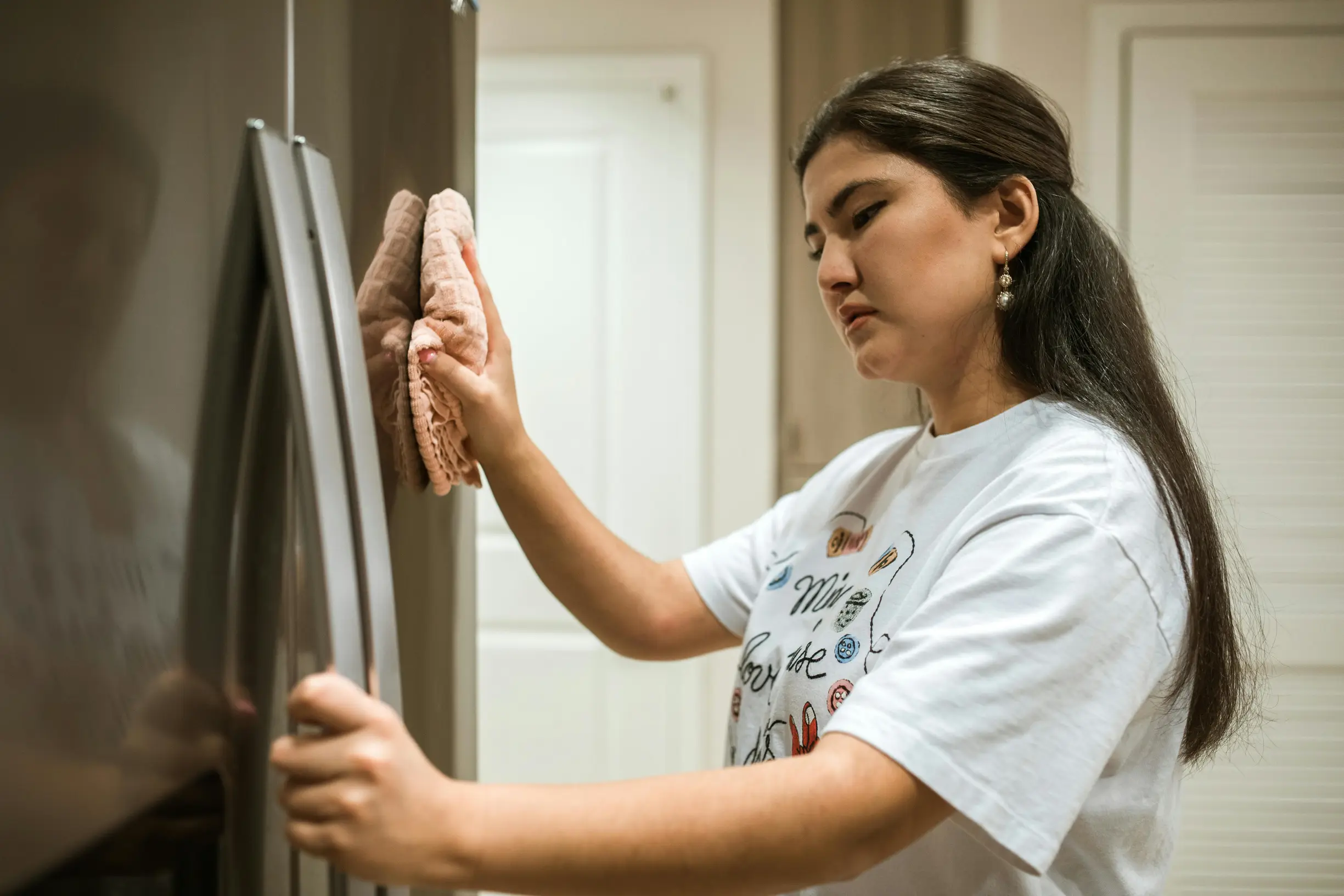 A young woman is cleaning the front of a stainless steel refrigerator with a cloth in a kitchen setting. She is standing sideways to the camera, slightly angled to the right, wearing a white graphic t-shirt. The kitchen appears indoors with a neutral-toned wall and door visible in the background. The lighting is soft and even, suggesting artificial indoor lighting, and the image is a medium shot showing the woman from about the waist up.