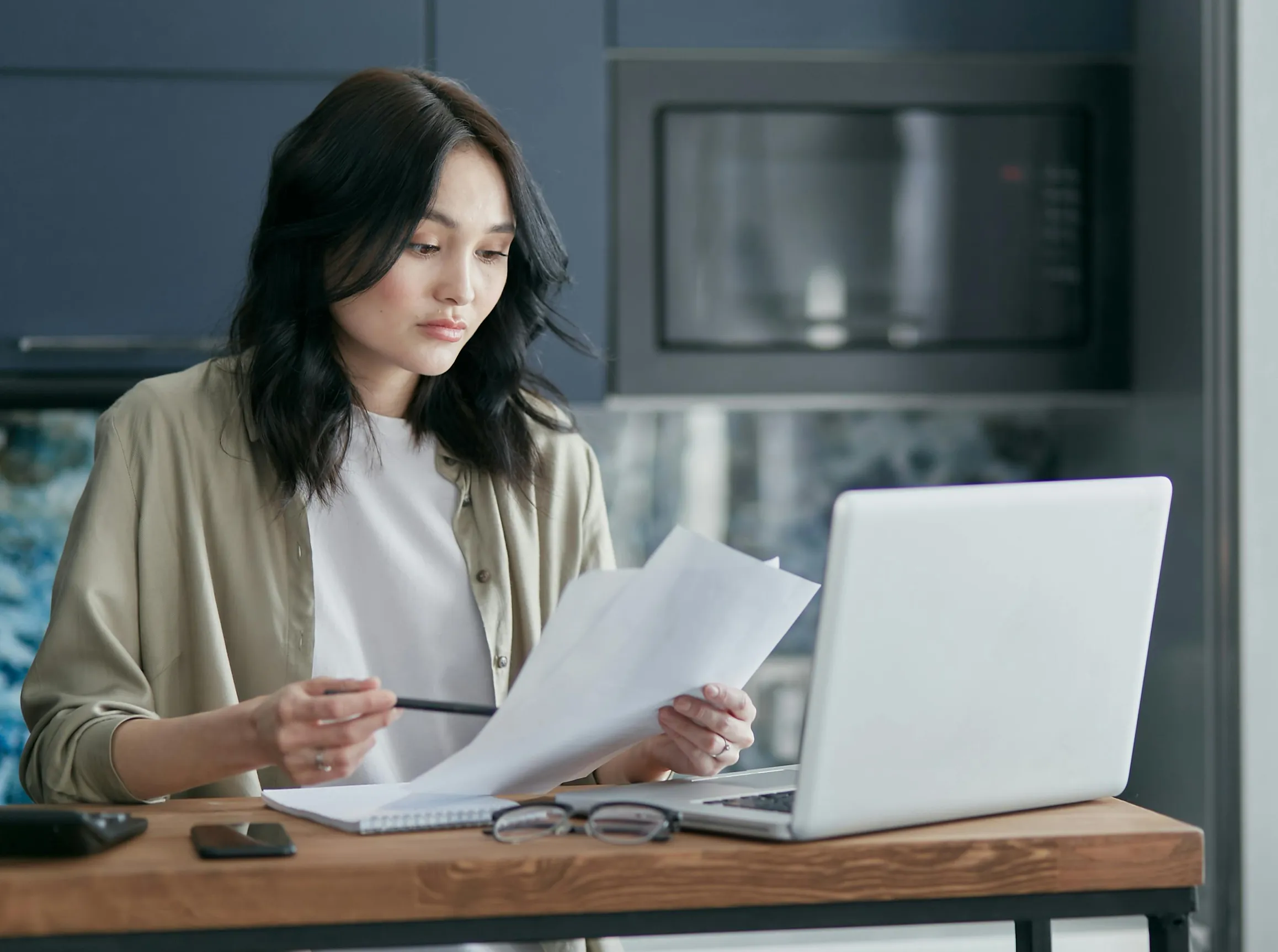 A young woman with medium-length dark hair is seated at a wooden table in what appears to be a kitchen or home office setting. She is wearing a light beige cardigan over a white top. In front of her is an open laptop, a notebook, a pair of eyeglasses, and a smartphone on the table. She holds several sheets of paper and a pencil in her hands, looking at the papers thoughtfully. Behind her is a microwave oven and a blue-gray wall with a textured backsplash. The lighting is soft and natural, with a neutral col