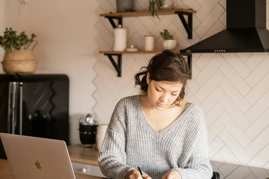 A young woman with medium-length dark hair tied back is seated at a kitchen island counter inside a modern kitchen. She is wearing a light gray knitted sweater and writing in a notebook with a pen. A laptop is open on the counter to her left. The kitchen features a black refrigerator, black range hood, white cabinets, and light wood surfaces. There are two black pendant lights hanging above the counter, and wooden shelves mounted on a white tiled wall hold some small plants and kitchen items.