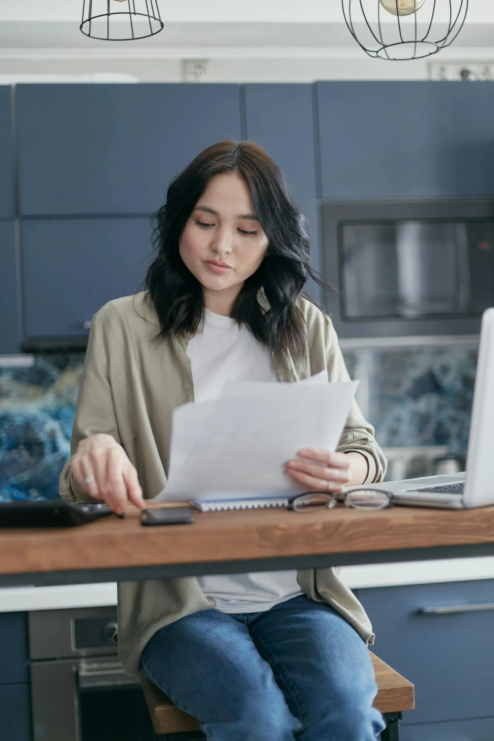 A young woman with shoulder-length dark hair is sitting at a wooden kitchen counter. She is looking down at several papers in her left hand while using a calculator with her right hand. The woman is wearing a beige overshirt over a white top and blue jeans. Behind her, there are blue kitchen cabinets, a microwave, and a modern backsplash. A laptop is open on the counter next to her. The scene is lit with natural or soft artificial light, and the photo is taken at eye level with a medium framing that capture
