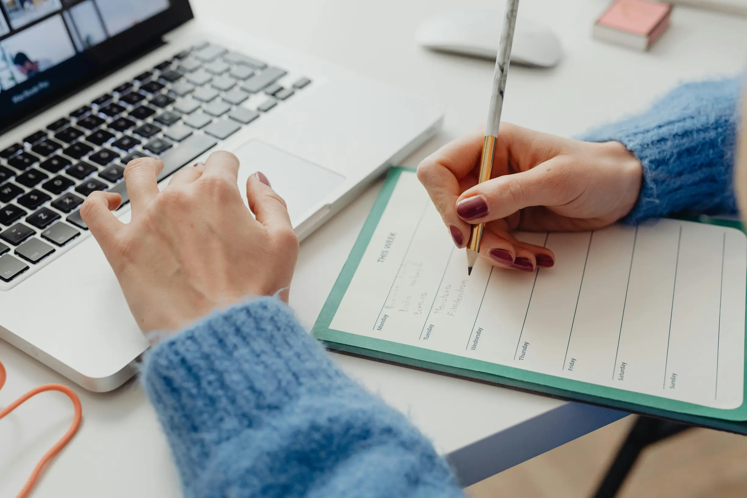 Image shows a close-up view of a person's hands working at a desk. One hand is typing or resting on a laptop keyboard, while the other hand is holding a pen and writing on a planner or calendar notebook. The person is wearing a blue knitted sweater. The desk surface is white, and a computer mouse and some small stationery items are visible in the background. The lighting is natural and evenly distributed, with a slight shadow cast by the hands and objects.