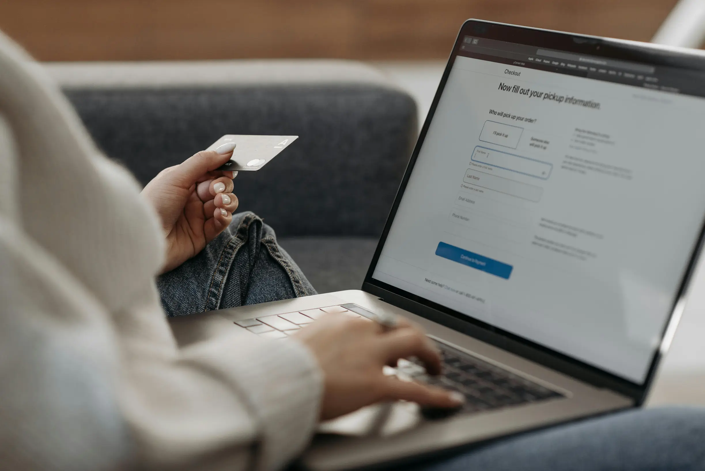 The image shows a close-up of a person using a laptop while holding a credit card. The person is seated on a gray couch, wearing a light-colored sweater and jeans. The laptop screen displays a web form requesting package information with fields labeled 'Where to?' and 'Who will receive this package?'. The person's left hand holds the credit card near the keyboard, and the right hand is poised on the laptop's keyboard or trackpad.