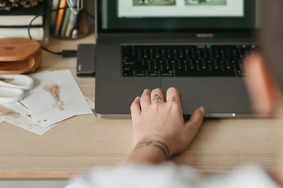 The image shows a close-up view from behind a person who is seated at a wooden desk working on a laptop. The person's right hand is on the laptop's trackpad or keyboard, but the individual's face is not visible. On the laptop screen, a grid layout of four photos displayed is visible. The desk has several items arranged including pencils in a holder, a pinecone, a large candle, a small cutting board, and scattered papers.