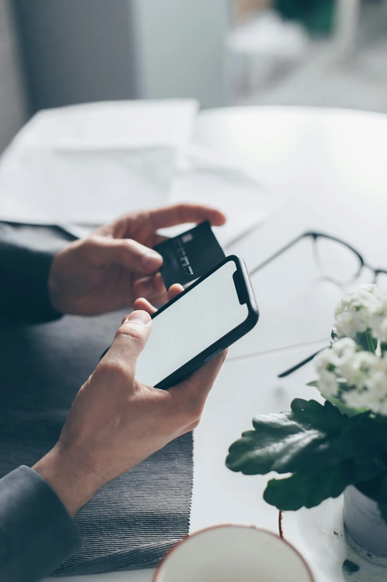 A close-up image of a person's hands holding a smartphone in one hand and a credit card in the other. The person is seated at a white table with a gray placemat. Nearby on the table are a pair of eyeglasses and a small potted plant with white flowers. Part of a light-colored cup is visible in the foreground. The background is softly blurred, suggesting a shallow depth of field.